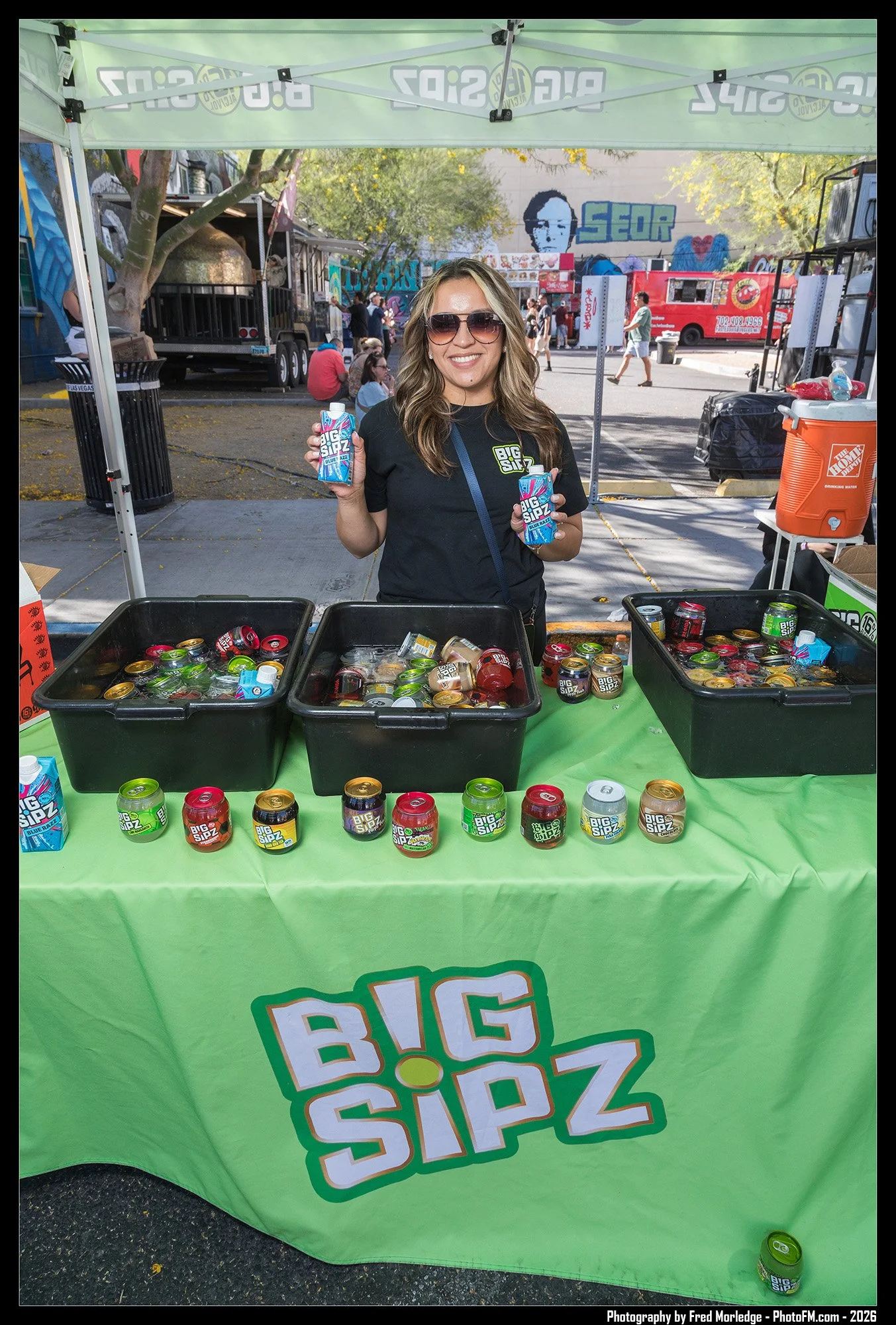 A smiling woman with sunglasses holding two juice boxes standing behind a table with cans of Big Sip drink at an outdoor event. There are other people, food trucks, and graffiti art in the background.