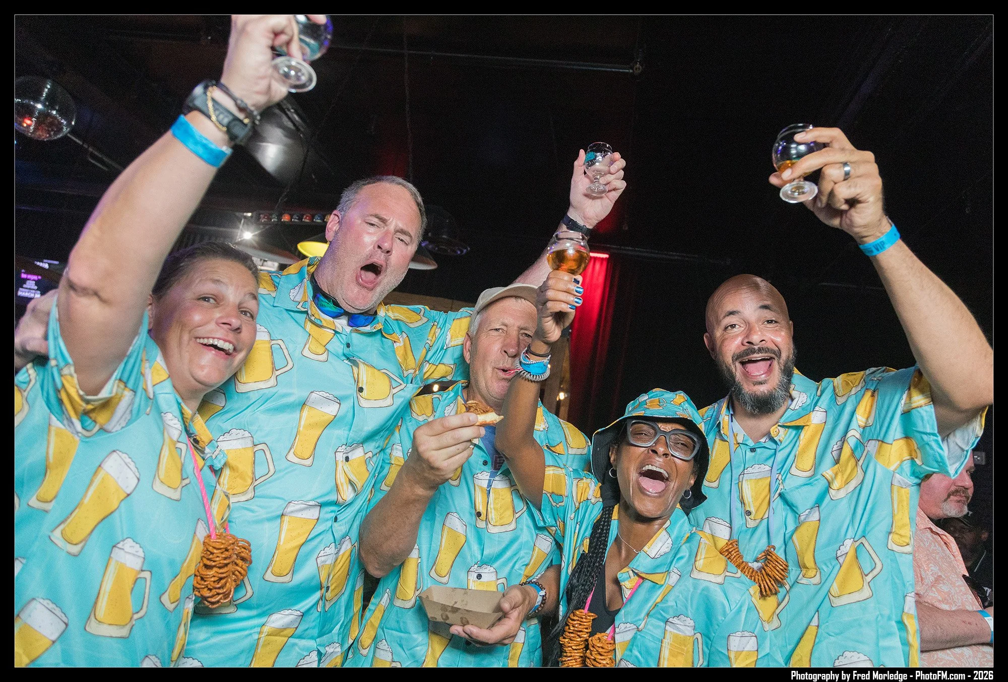 Group of friends celebrating in a bar, wearing matching shirts with beer and pretzel prints, raising glasses of beer, smiling, and enjoying the party.