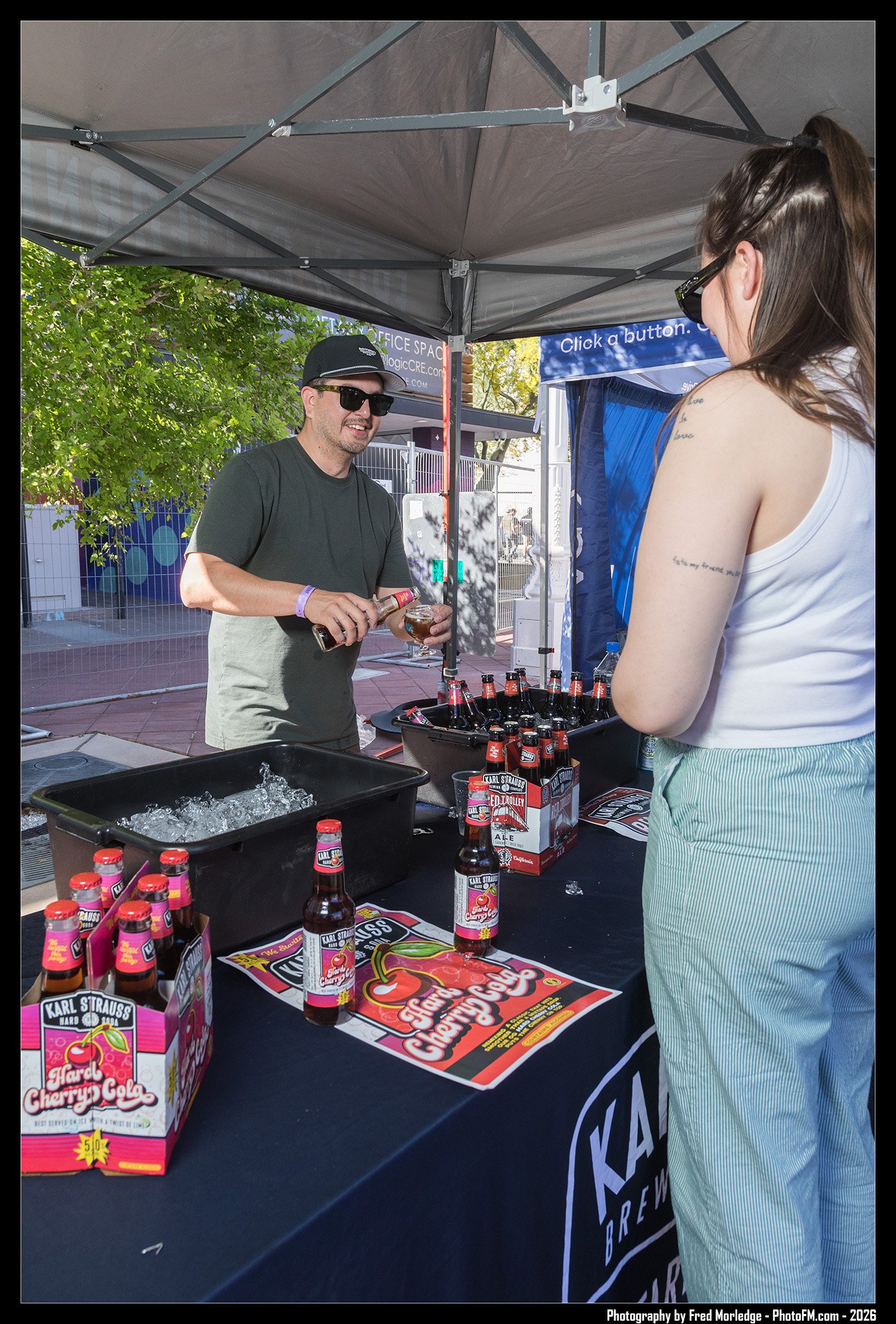 A man wearing sunglasses, a black cap, and a gray shirt is pouring a bottled drink into a glass at an outdoor event booth. A woman with tattoos on her arm, wearing a white tank top and sunglasses, faces him on the other side of the booth. The booth d