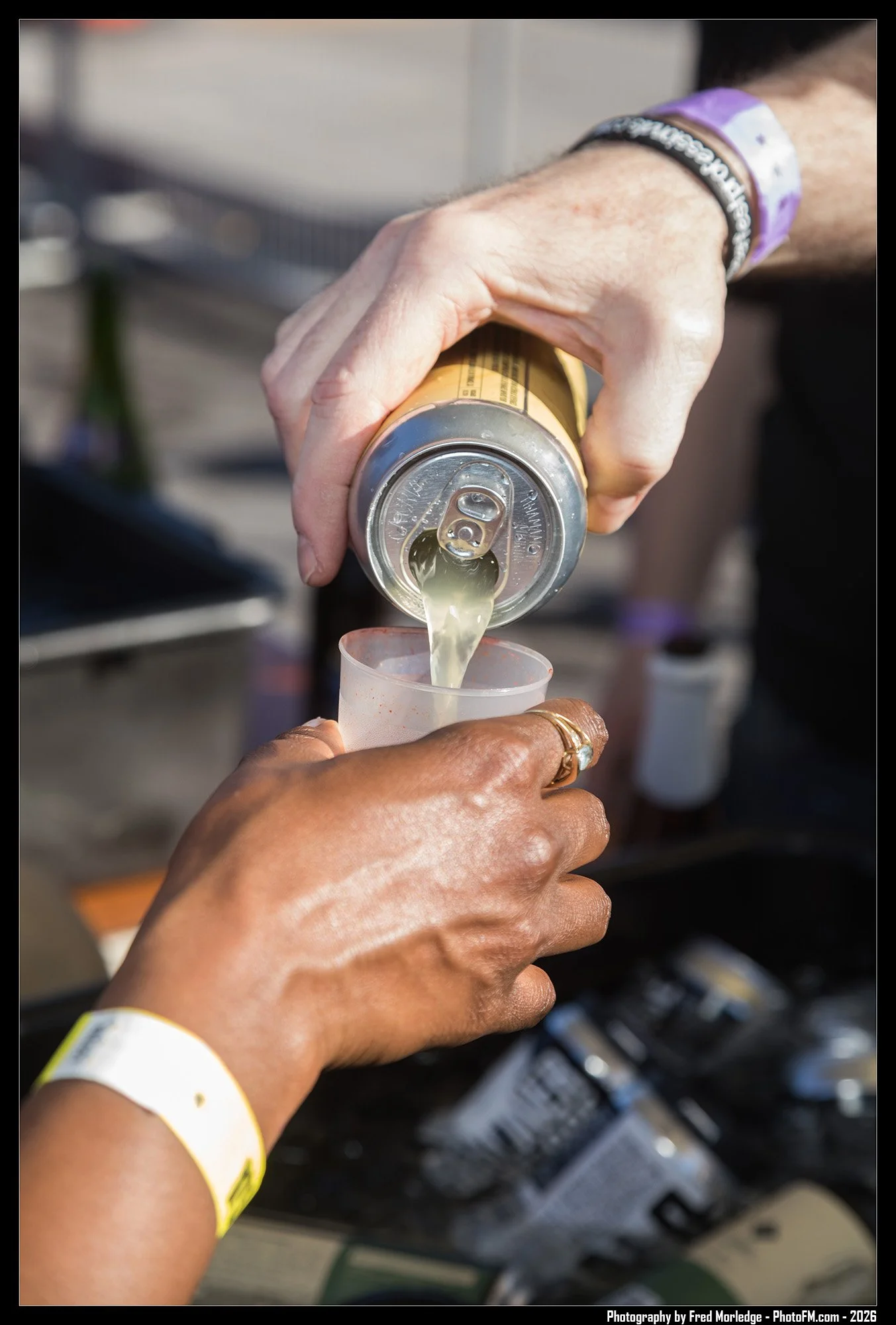 A person's hand is pouring a light-colored beverage from a can into a clear plastic cup. The person is wearing a yellow wristband and has a ring on their finger. The background shows part of a vending machine.