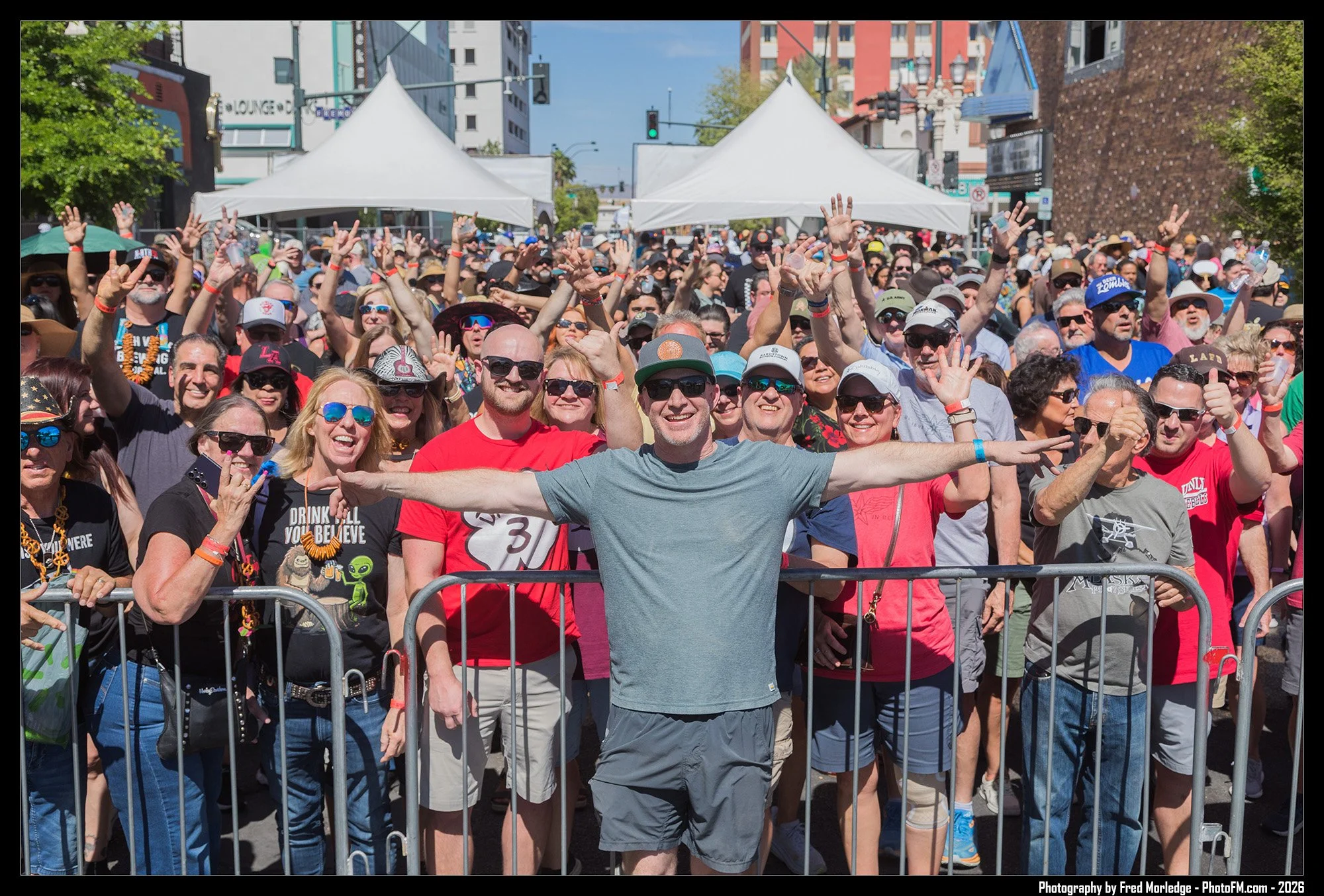 A large crowd of people gathered outdoors in front of a stage, with many smiling and raising their hands, at a daytime event on a city street, under blue skies with tents and buildings visible in the background.