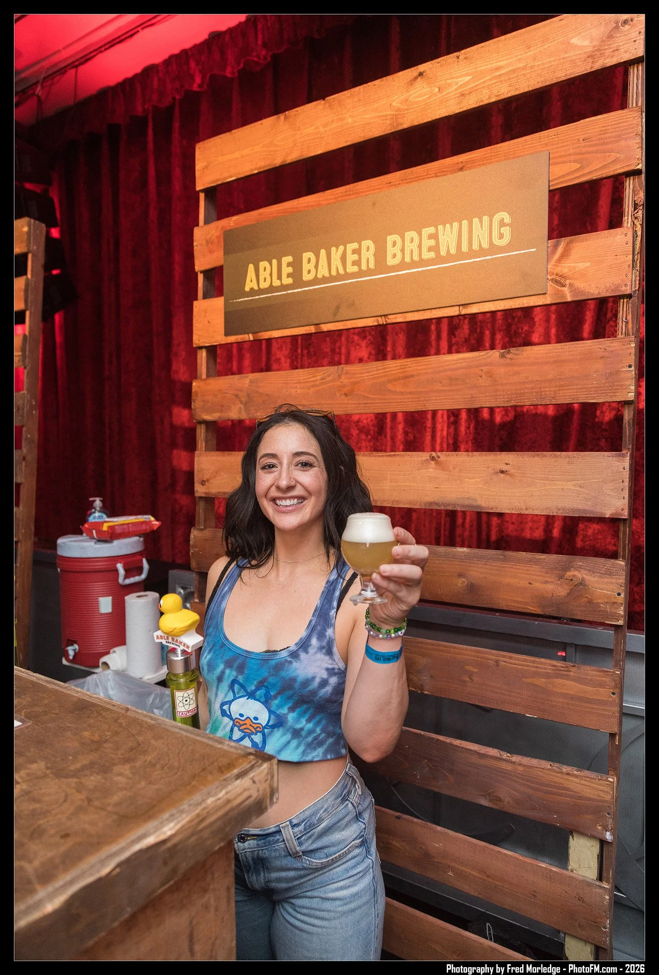 A woman with dark hair and a big smile, wearing a tie-dye tank top with a cartoon character on it, is holding a glass of beer at the Able Baker Brewing event. She has bracelets on her left wrist and is standing in front of a wooden sign with the brew
