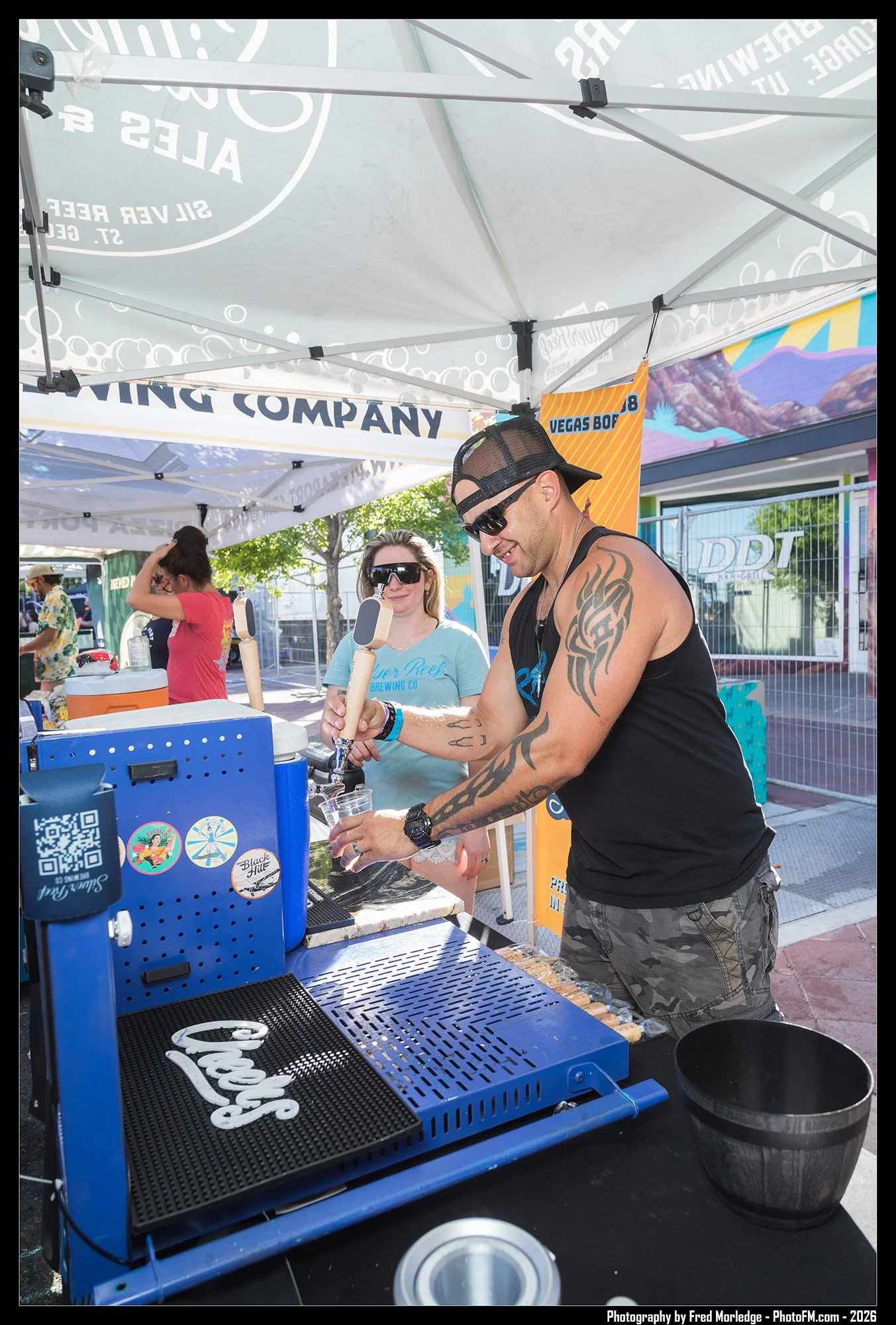A man with tattoos on hisarm, wearing a black tank top, camouflage shorts, sunglasses and a backward baseball cap, is using a blue beverage dispenser at an outdoor event. A woman with blond hair, wearing sunglasses and a light blue T-shirt, is standi