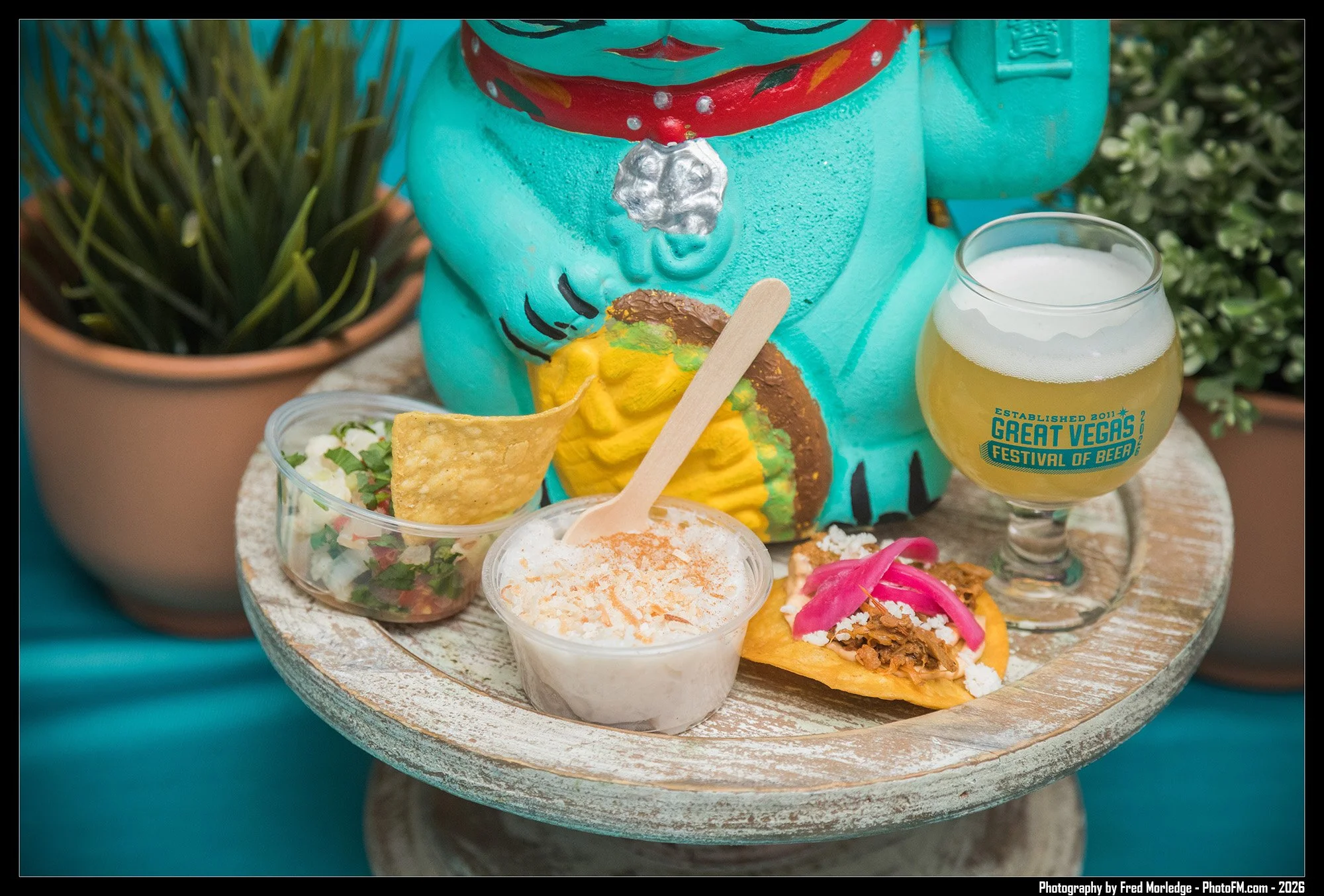 A collection of Mexican food and drinks on a round tray, including rice, salsa, chips, and tacos, with a teal-colored festive bear figure and a glass of beer on the side, set outdoors.