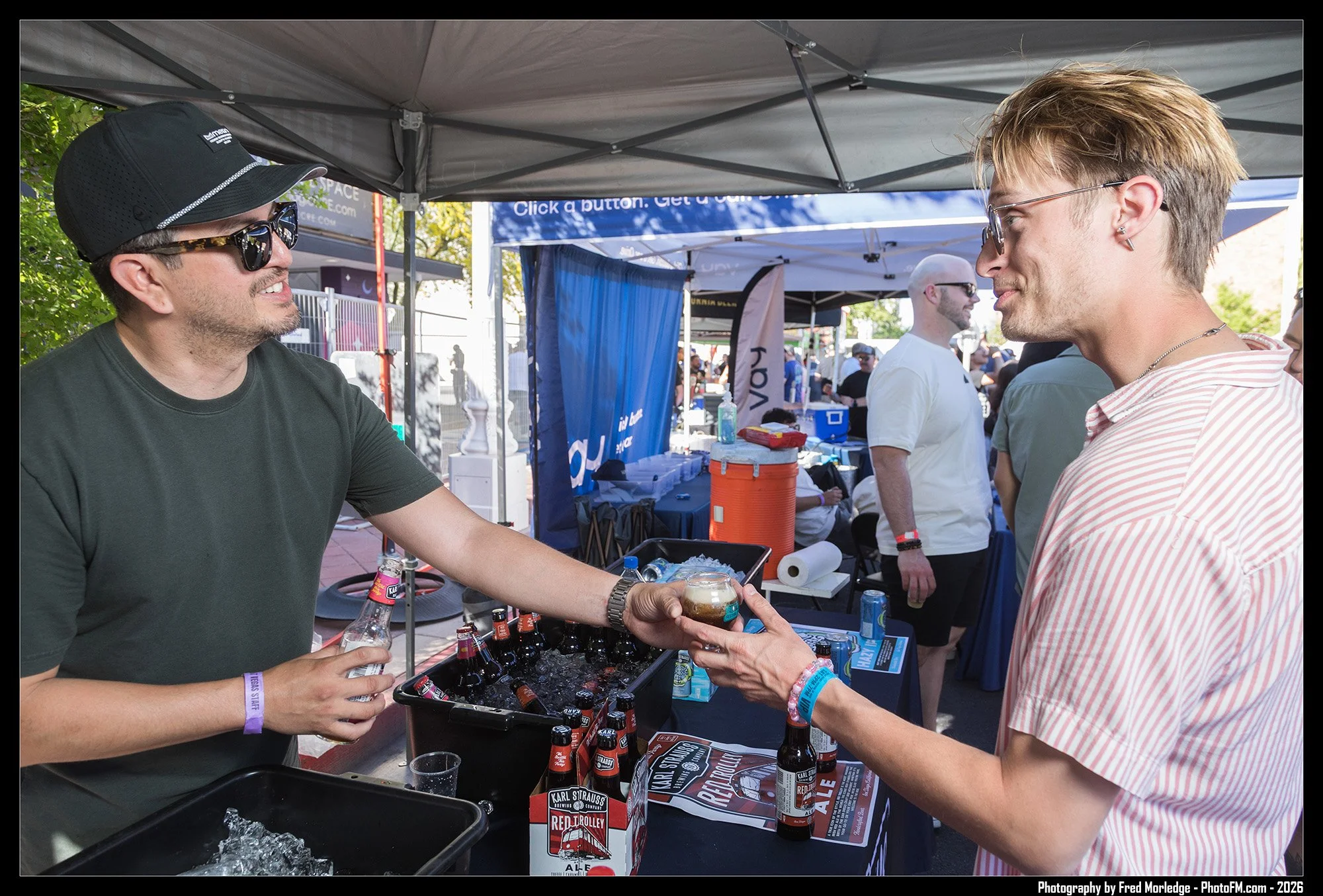 Two men exchanging drinks at an outdoor event, with tents and booths visible in the background, and bottles of beer and various items on the table between them.