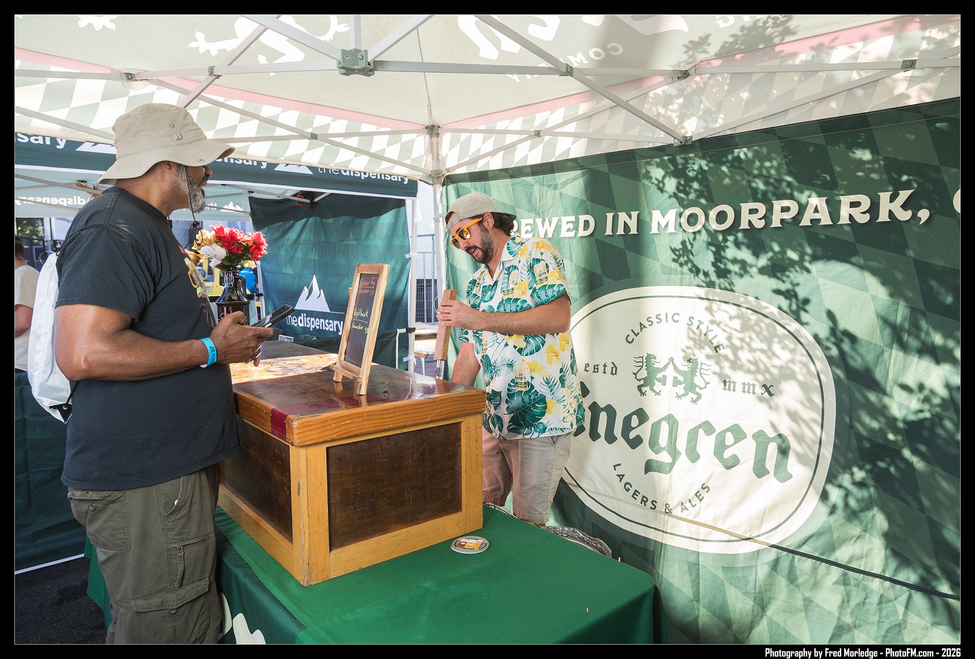 Two men at an outdoor booth, one wearing a beige sun hat and sunglasses, and the other wearing a cap and tropical shirt, standing in front of a green banner that reads 'Brewed in Moorpark, California, with a crest and text about the brewery.