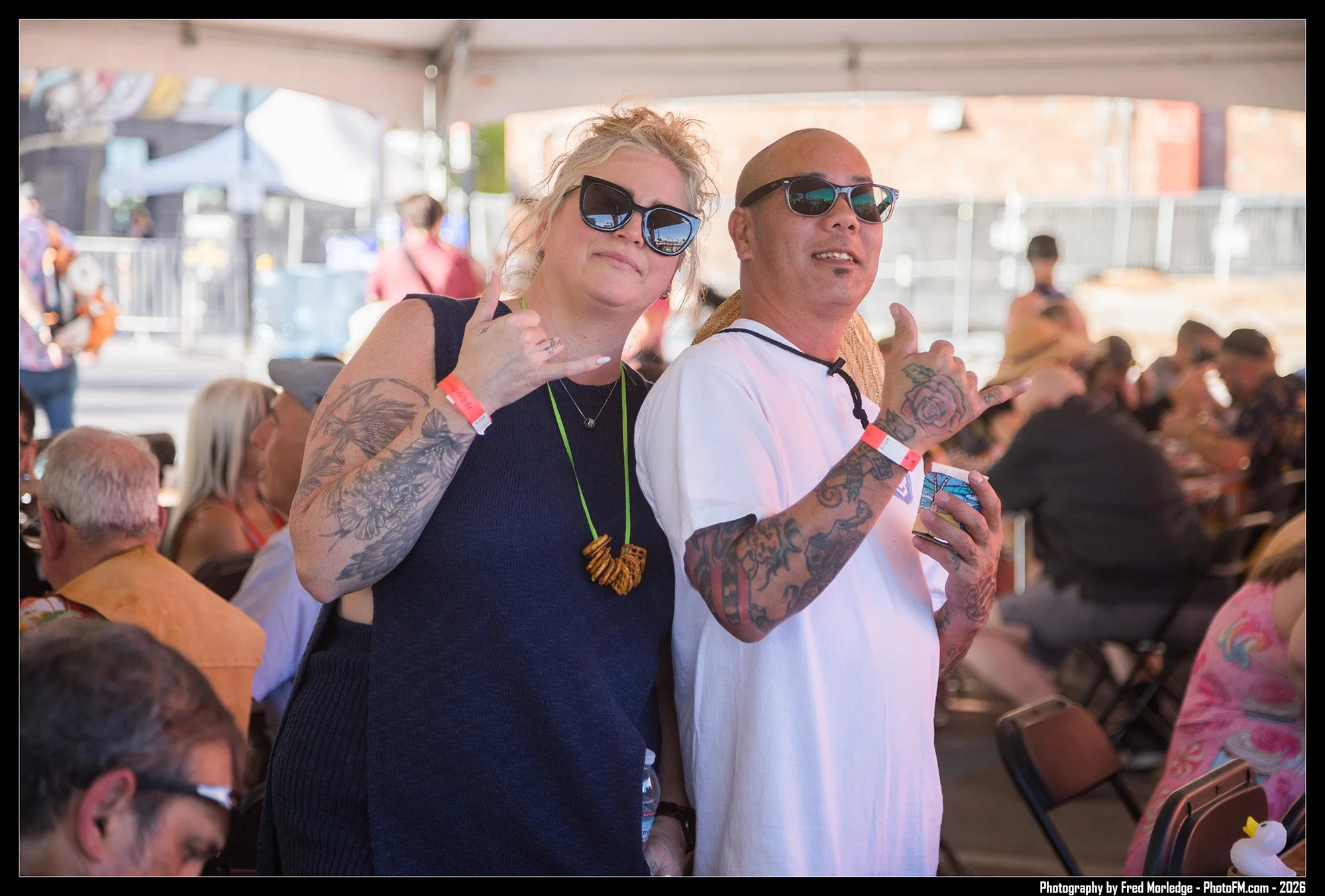Two people with tattoos and sunglasses making the shaka hand gesture at an outdoor event, with many people sitting at tables under a tent.