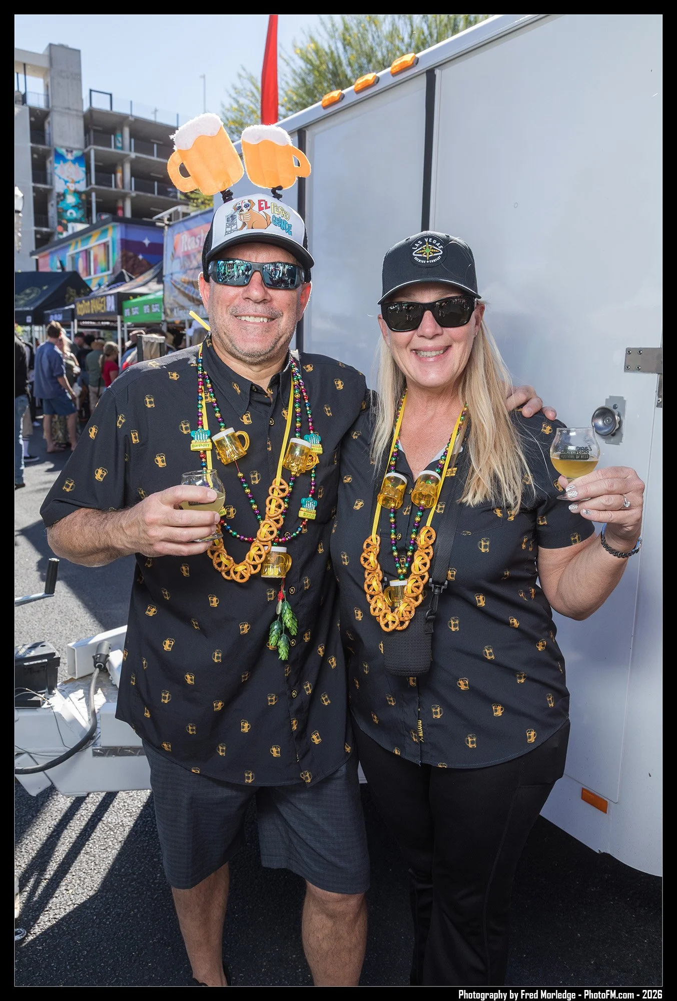 Two people smiling at a festival, wearing matching shirts with beer mug patterns and festive necklaces. The man on the left is wearing sunglasses and a hat with beer mugs and a cartoon animal, with plush beer mugs on his head. The woman on the right 