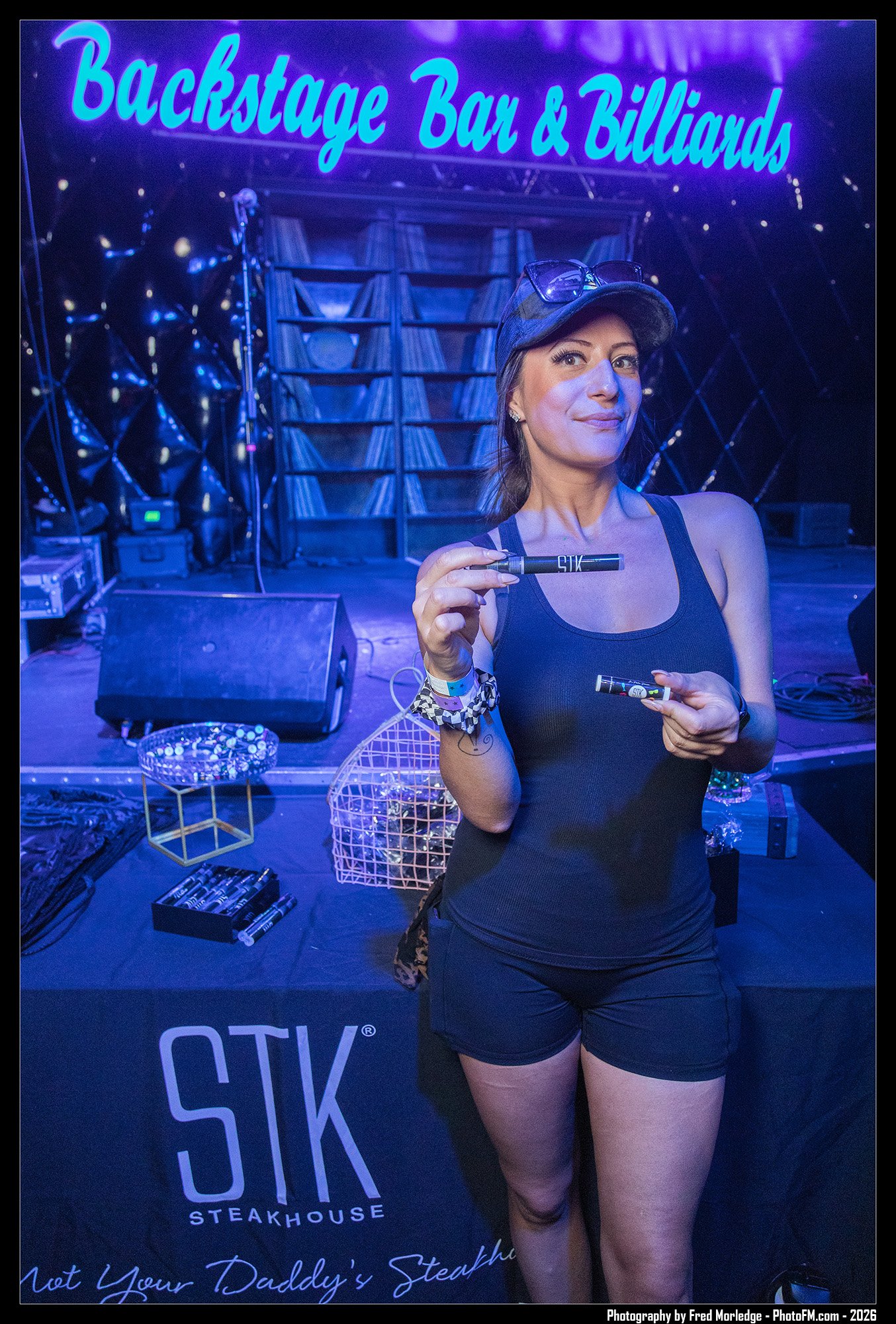 A woman in athletic attire holding a marker inside a neon-lit stage area with a backdrop that reads "Backstage Bar & Billiards" and "SIX Steakhouse."