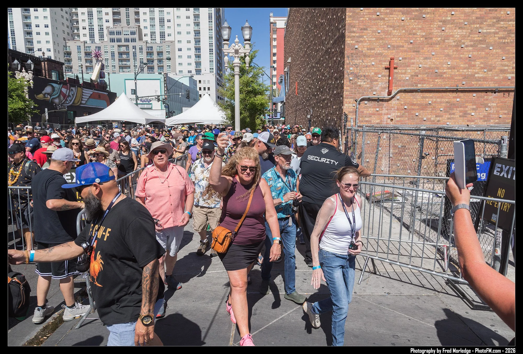 Crowd of people walking outdoors in an urban area during daytime, with some tents and tall buildings in the background.