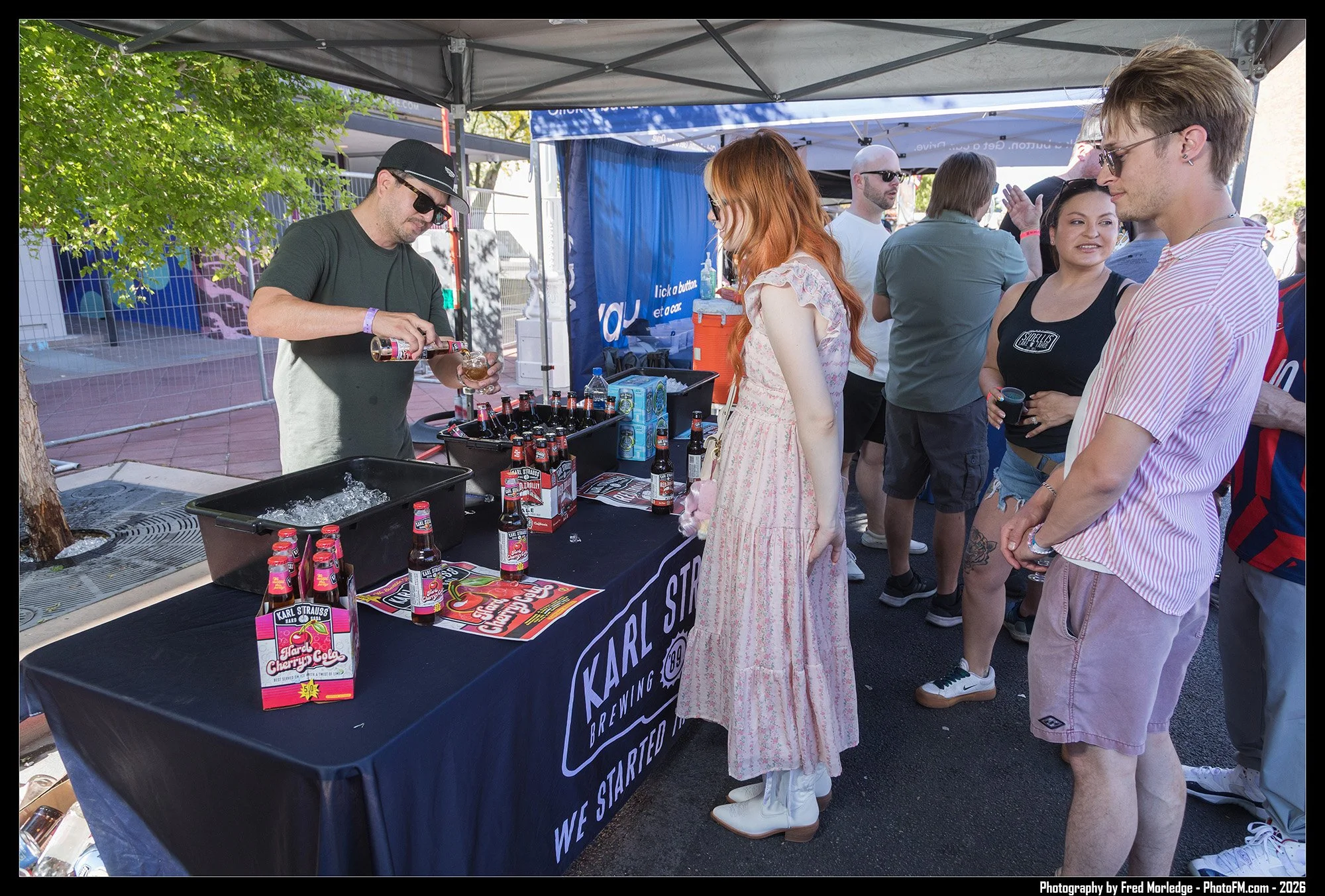 A group of people at an outdoor event at a Karl Strauss Brewing Company booth. A man is pouring a drink for a woman with red hair, while others wait in line or socialize nearby. There are bottles of Karl Strauss Cherry Cola on the table.
