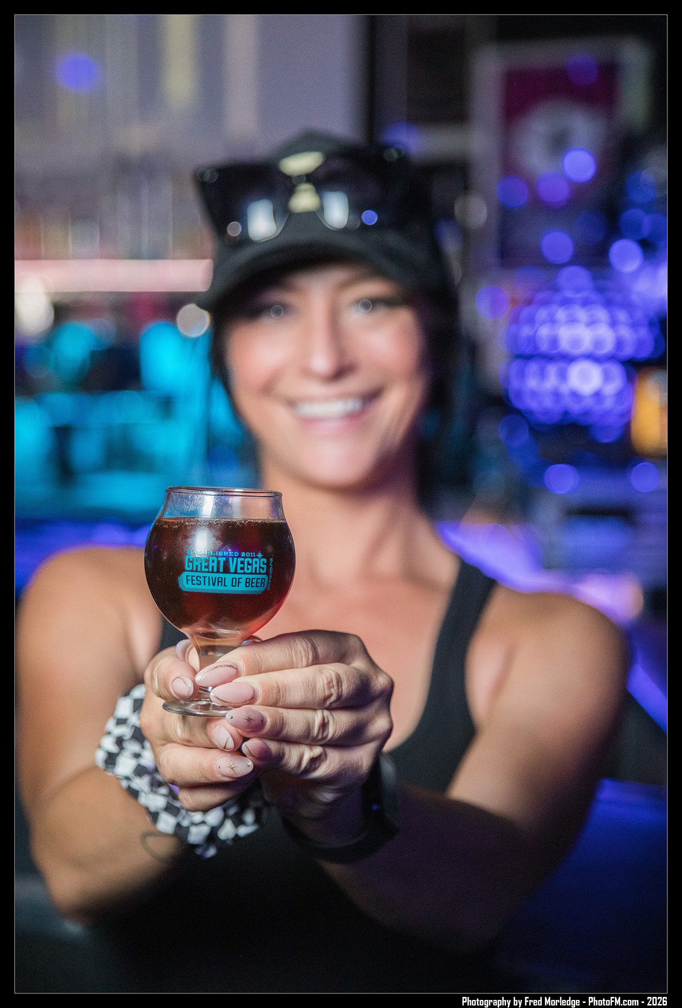 A smiling woman in a black tank top, wearing glasses on her head, holds a glass of dark beer with a label that says 'Great Vegas Festival of Beer' at a nighttime event with colorful bokeh lights in the background.