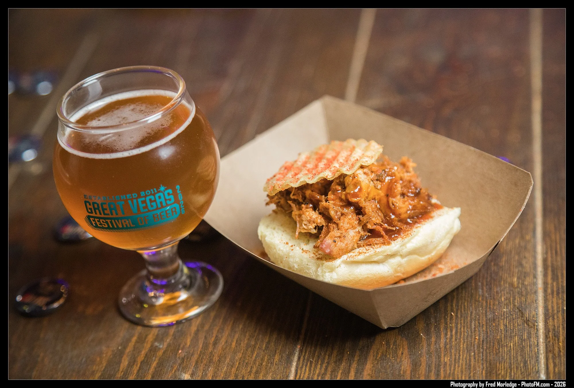A glass of beer with a logo reading 'Great Vegas Festival of Beer' and a sandwich with shredded meat on a bun, served on a paper tray on a wooden table.
