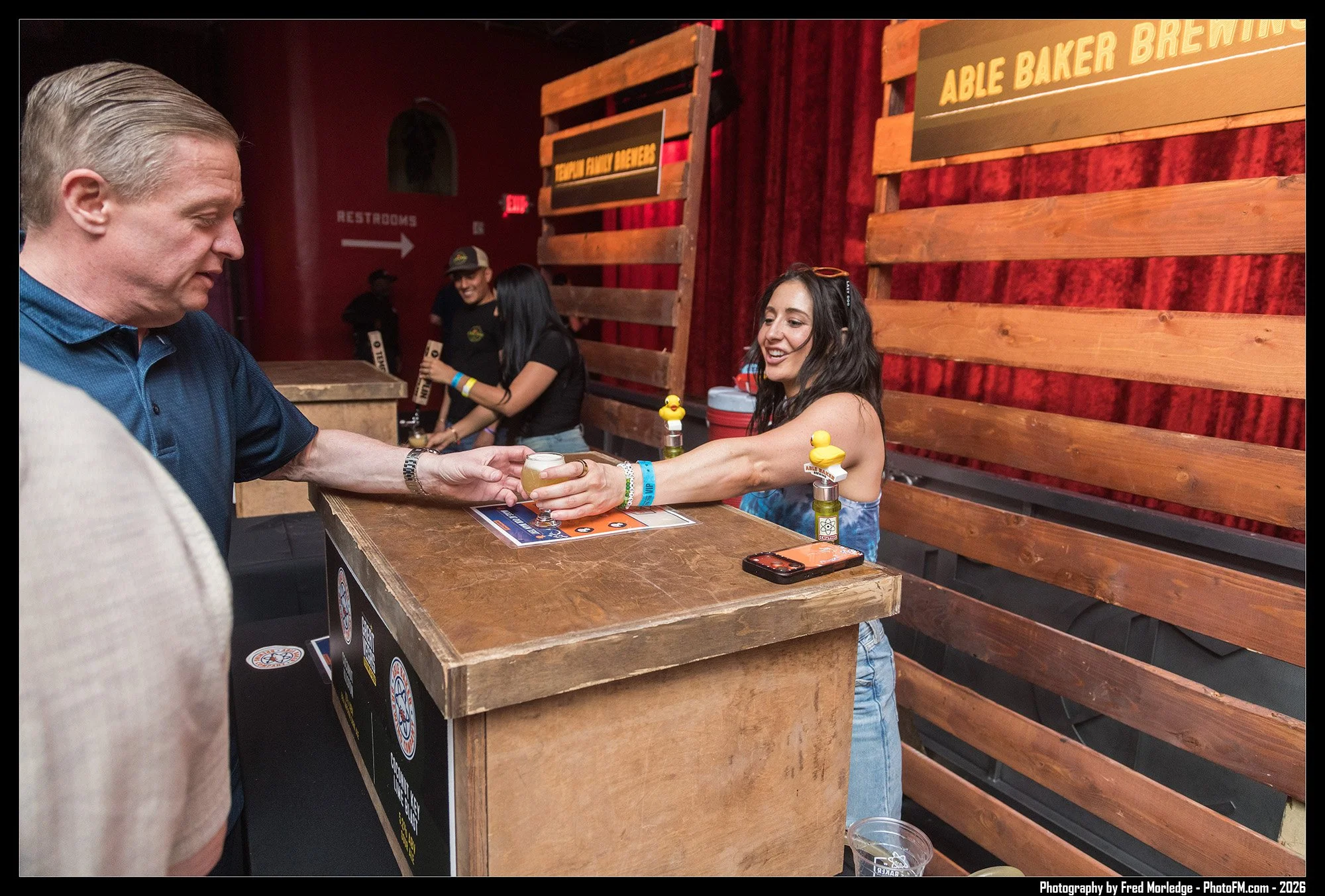 A young woman with long dark hair serving a drink to a man at a booth at Able Baked Brewing, with other people in the background. Wooden signs and red curtains decorate the background.
