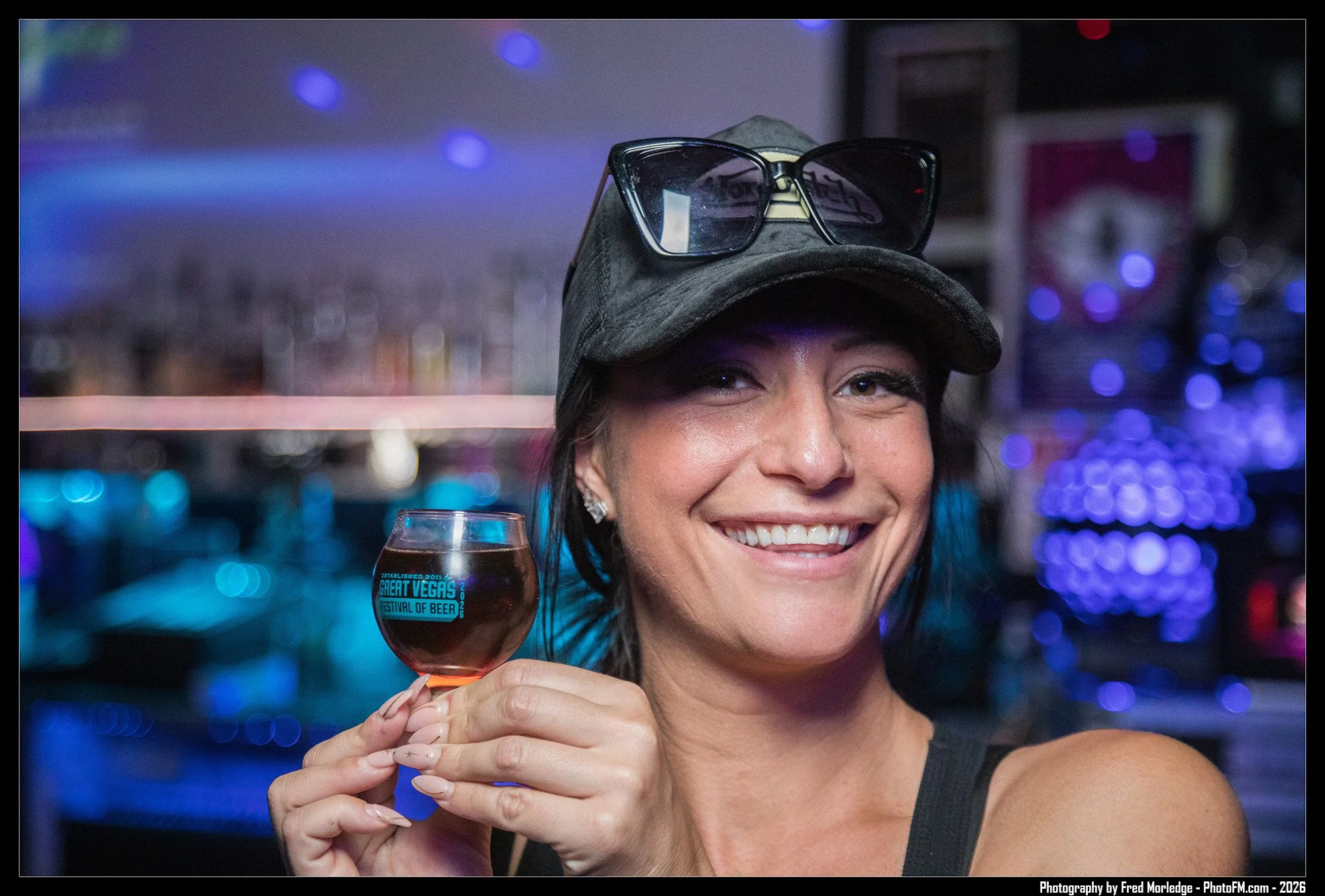 A woman smiling and holding a glass of dark beer, wearing sunglasses on top of her head and a casual black top, with a blurred colorful background featuring lights.