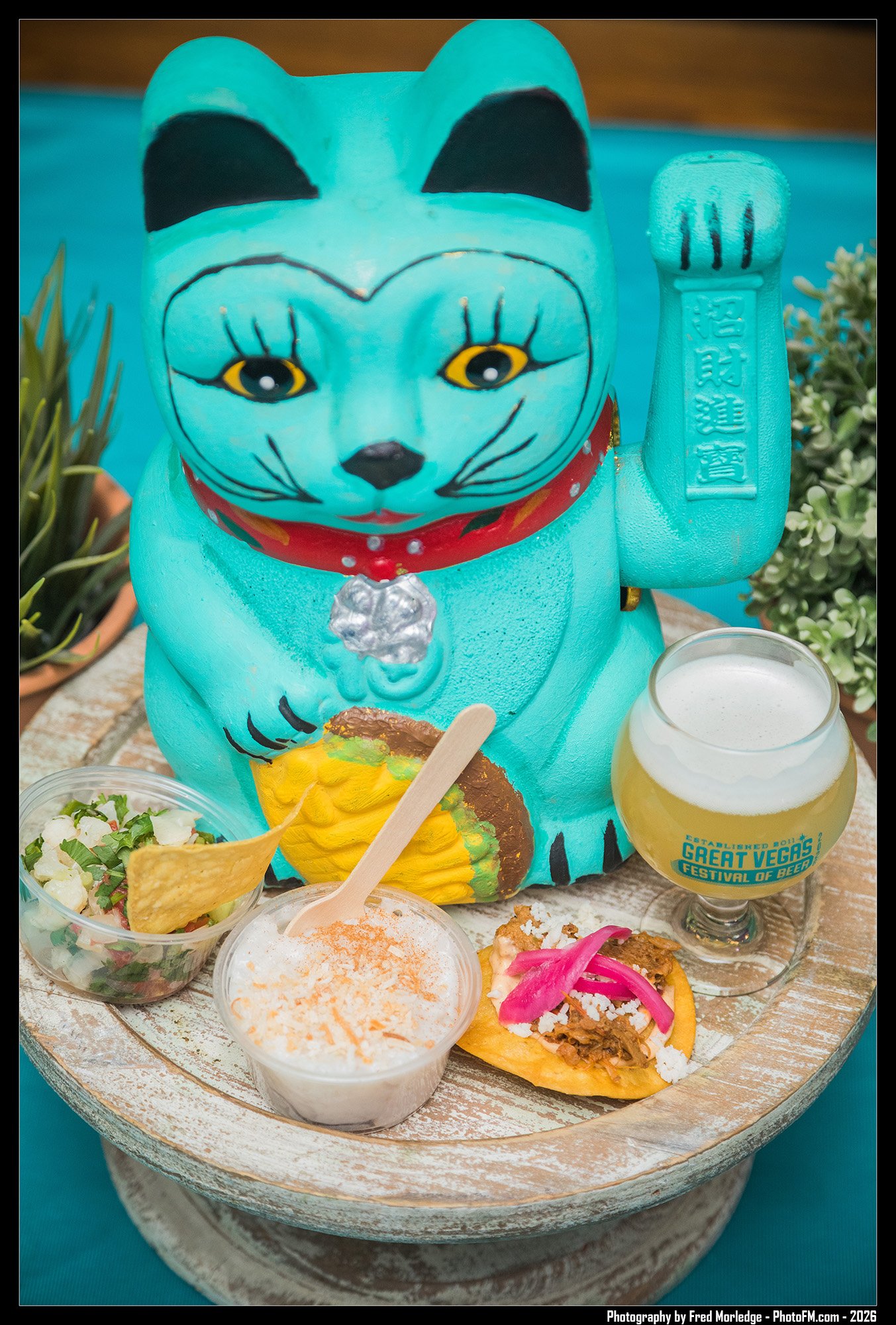 A Maneki-neko figurine with a light blue body, black eyes, yellow accents, and a red collar, surrounded by small food portions, including a bowl of salad, a creamy dip, a beer glass, and tacos, on a wooden tray with green plants in the background.