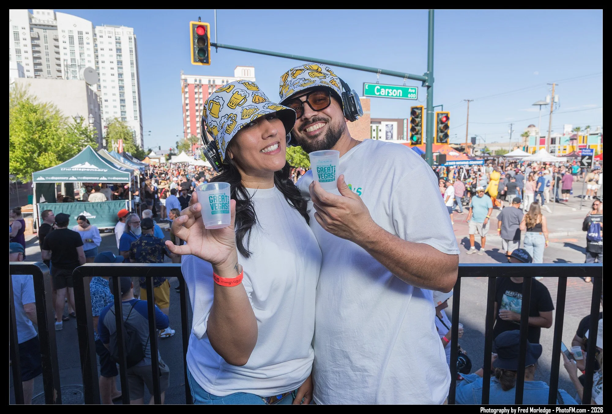 Two people wearing matching hats with beer mug pattern, holding cups with 'Great Vegas!' written on them, at an outdoor event in Las Vegas on Carson Avenue.