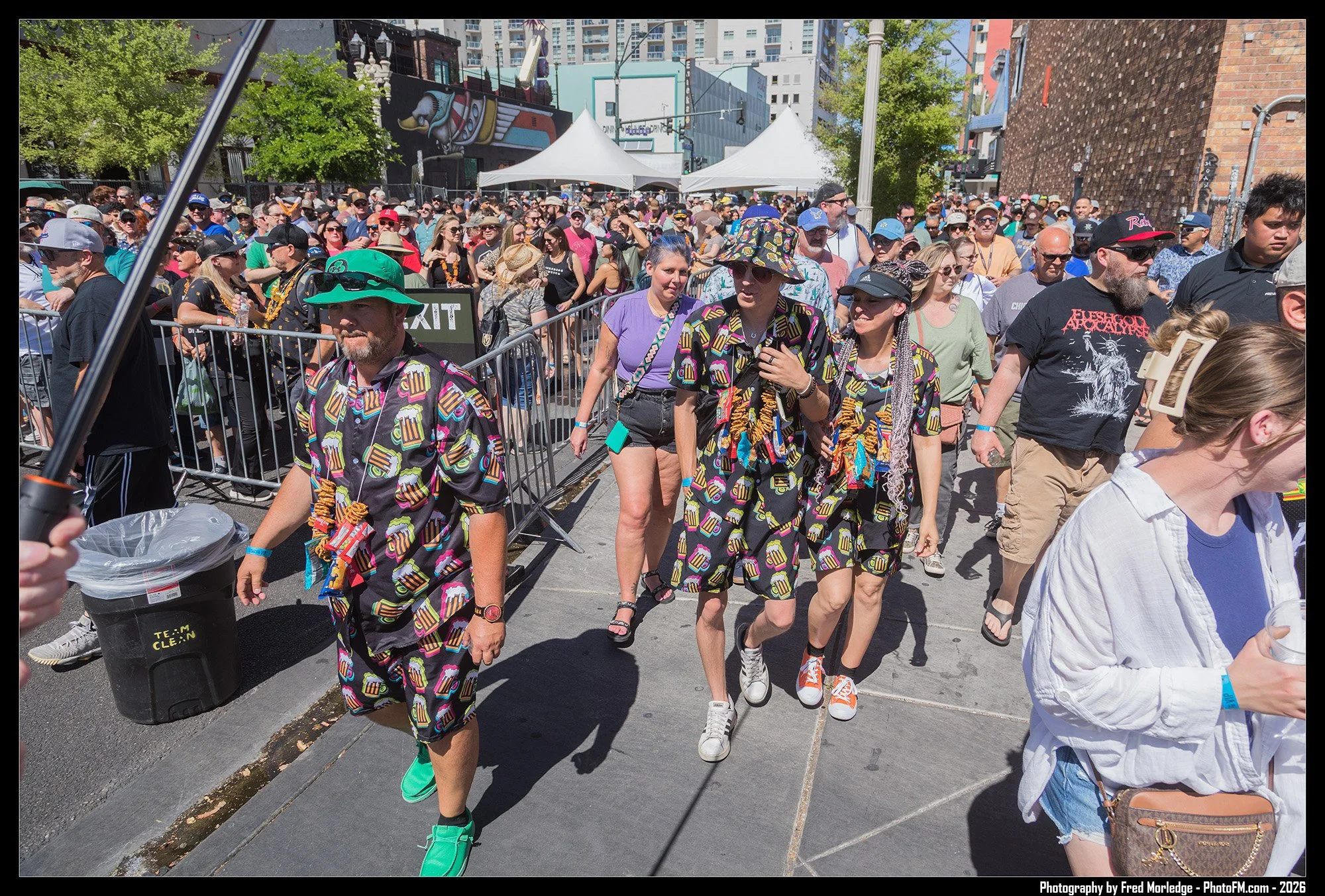Crowd of people walking outdoors at a festival or event, some wearing colorful clothing and hats, with tents in the background and tall buildings.