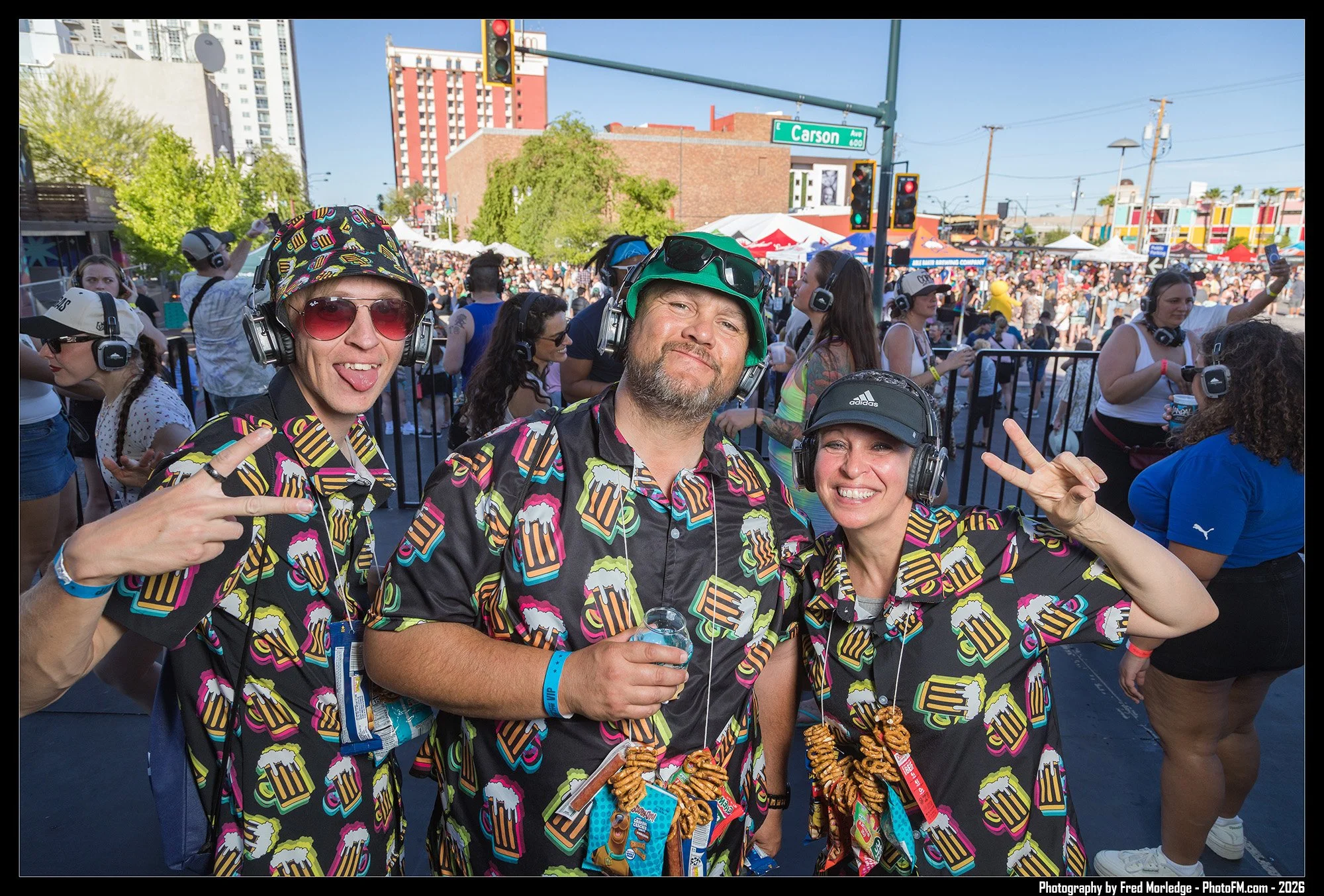 Three people at an outdoor event, wearing colorful shirts with ice cream patterns, headphones, and posing for a selfie. The background shows a large crowd, tents, trees, and a city street with a traffic light and street signs.
