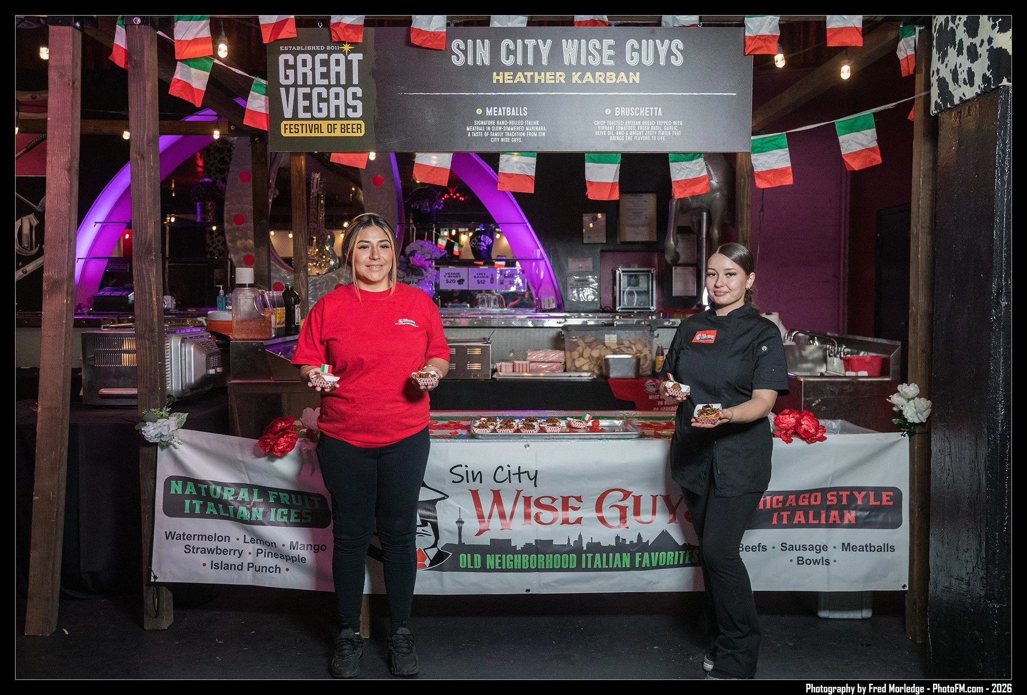 Two women standing in front of a food stand at the Great Vegas Festival of Beer, with a sign that reads 'Sin City Wise Guys' Heather Karban, offering Italian meats, meatballs, bruschetta, and Italian-style foods. The stand is decorated with Italian f