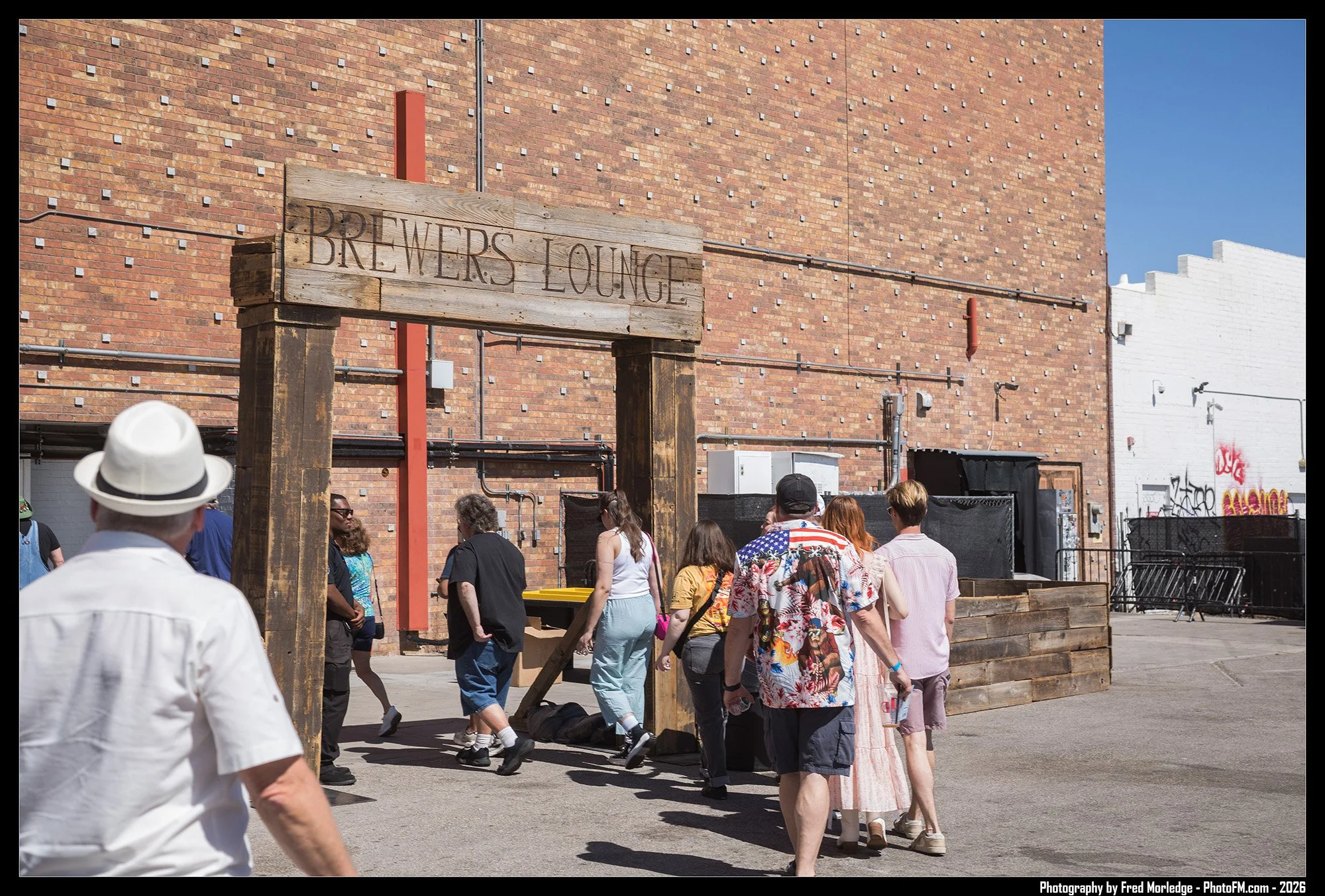 A group of people walking past a brick building with a wooden sign that reads "BREWER'S LOUNGE". The scene is in an urban outdoor area with a mix of individuals dressed casually, including some in summer clothing. There is graffiti on the white wall 