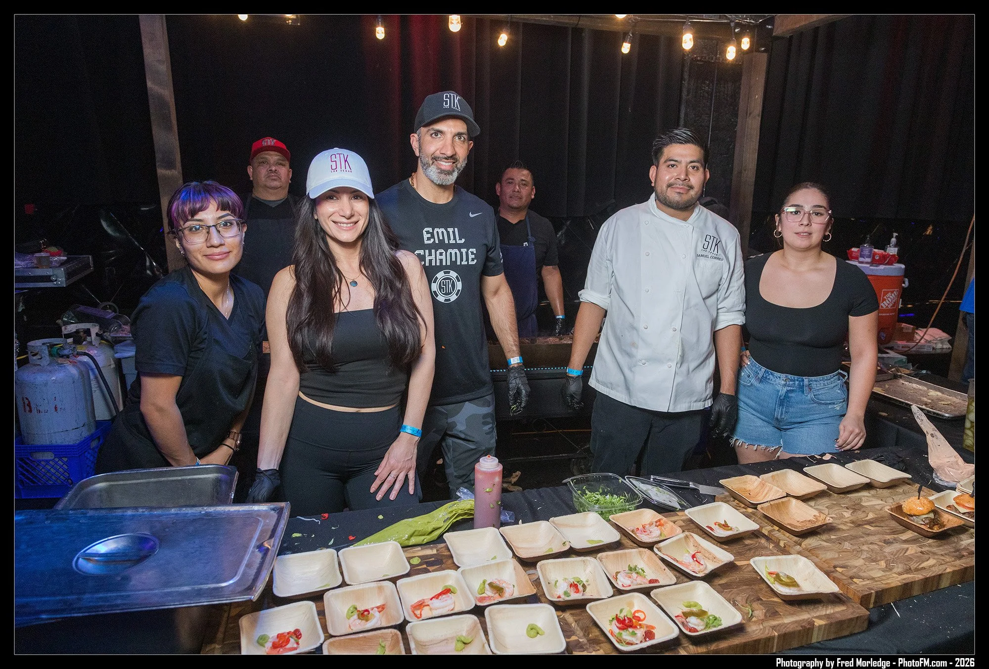People standing behind a table with food trays at a restaurant or event. The group includes four women and three men, with some wearing black aprons, chef jackets, and hats. The table has various food items and condiments.