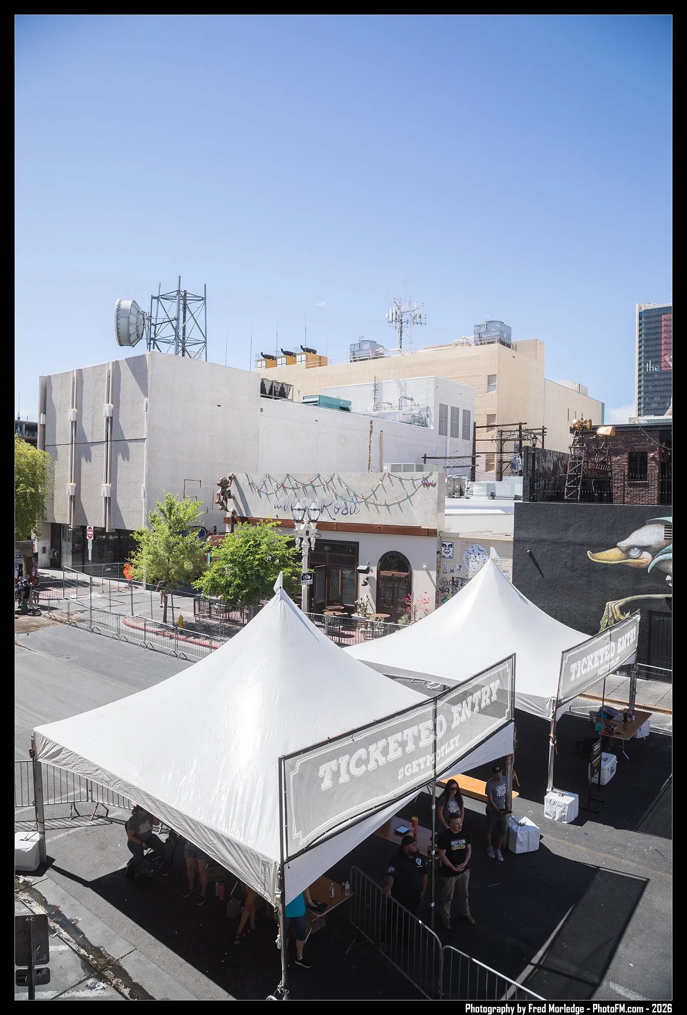 Outdoor event with white tents, people, and barricades, set against city buildings with graffiti and communication antennas on rooftops under a clear blue sky.