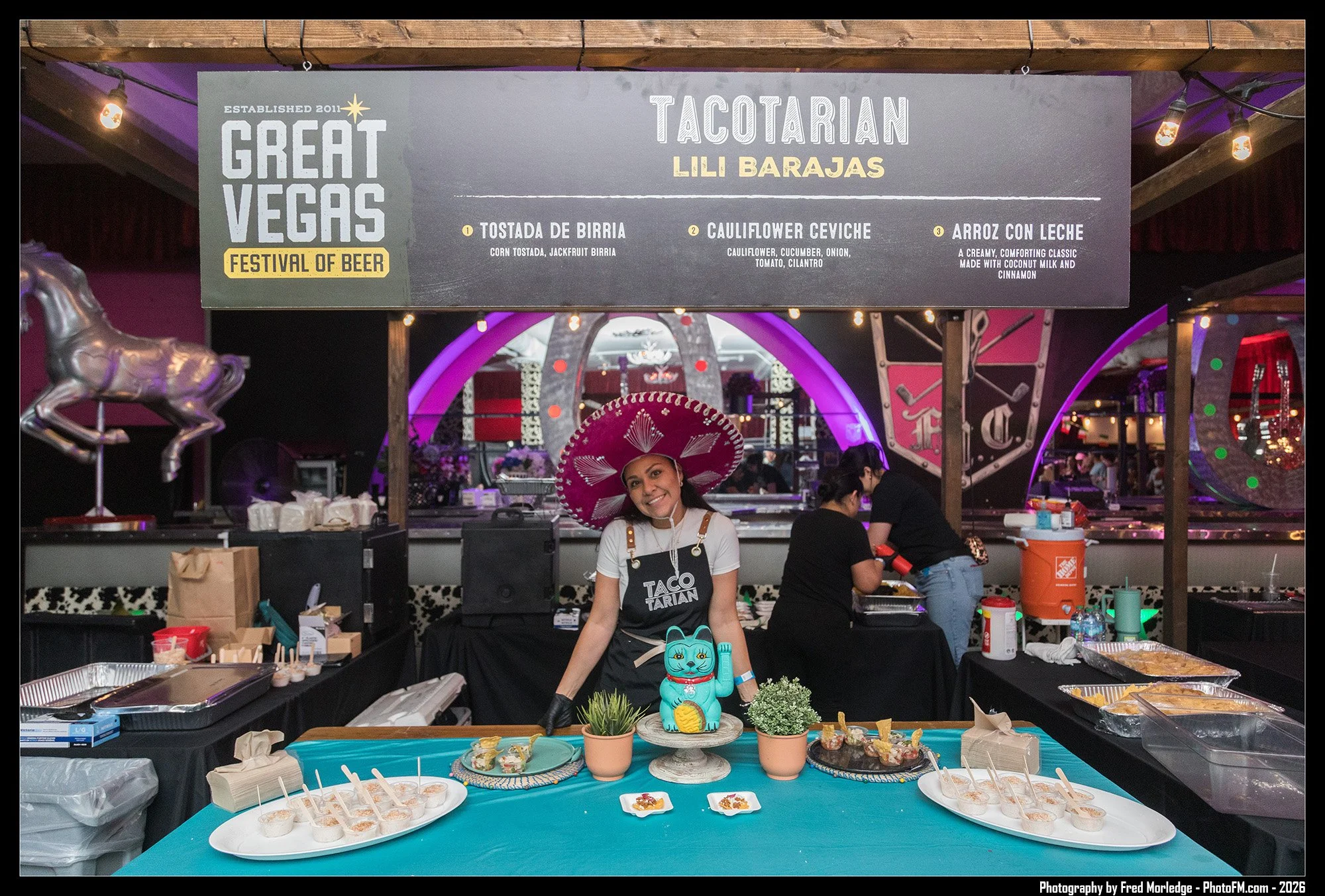 Food vendor at a festival serving Mexican-inspired dishes, including a woman wearing a large pink sombrero and a black apron with the words "Taco Tarian", surrounded by dishes of food and small potted plants, with a vibrant background and festival si