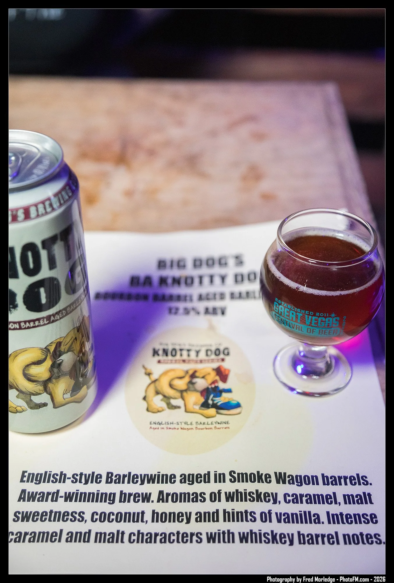 A can of English-style barleywine beer, a glass of dark beer with branding for the Great Vegas Festival of Beer, and a paper with an illustration of a cartoon lion and a man, along with the text "English-style Barleywine aged in Smoke Wagon barrels."