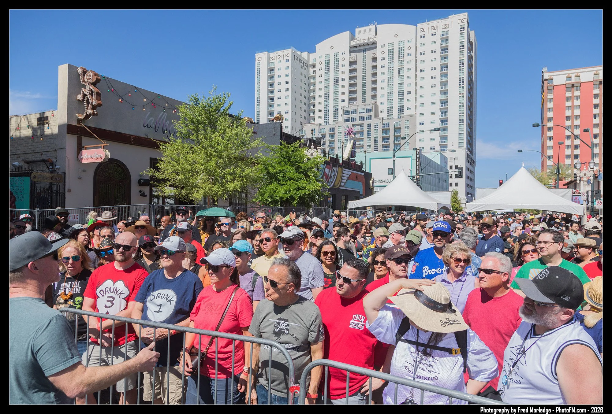 Crowd of people gathered outdoors at a festival or protest, with some wearing hats, sunglasses, and t-shirts with slogans, and standing behind a metal barrier on a sunny day, with tall buildings and tents in the background.