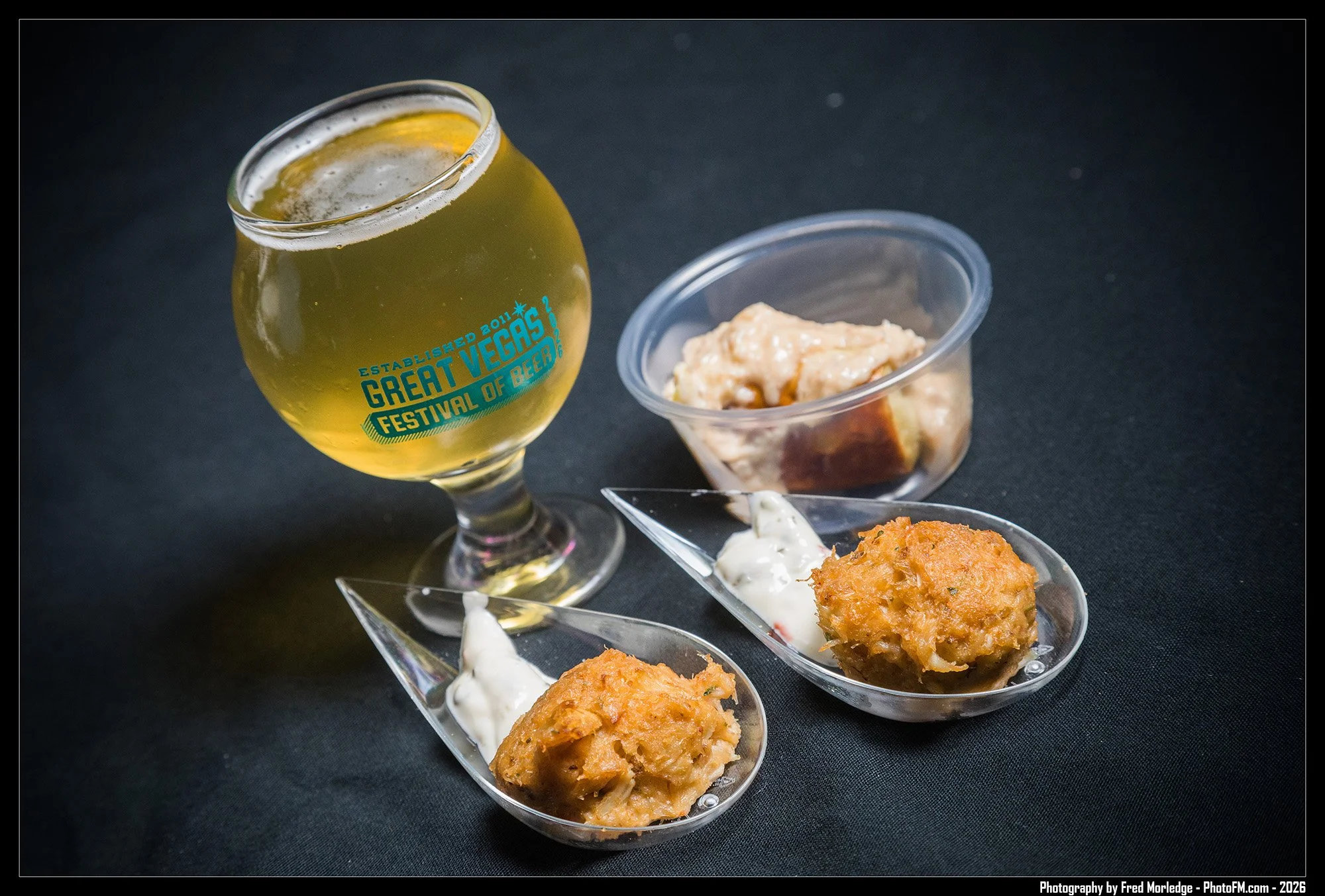 A glass of light-colored beer with a blue logo that reads, "ESTABLISHED 2011 GREAT VEGAS FESTIVAL OF BEER" sitting next to a small bowl of creamy dip. In front, on a black surface, are two small, clear holders filled with fried appetizers, possibly c