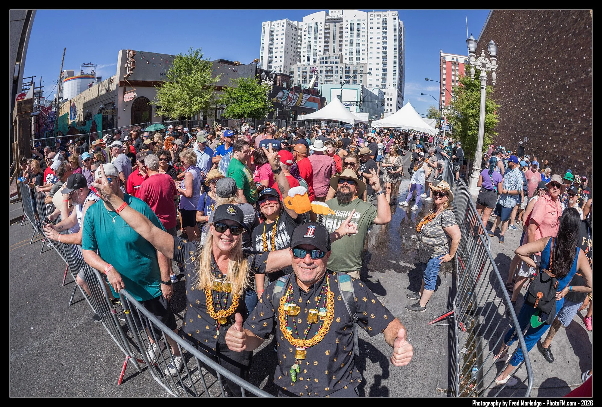 Crowd gathered at an outdoor event, people wearing colorful clothes, hats, and beads, with some holding drinks, in a city street with tall buildings and murals.