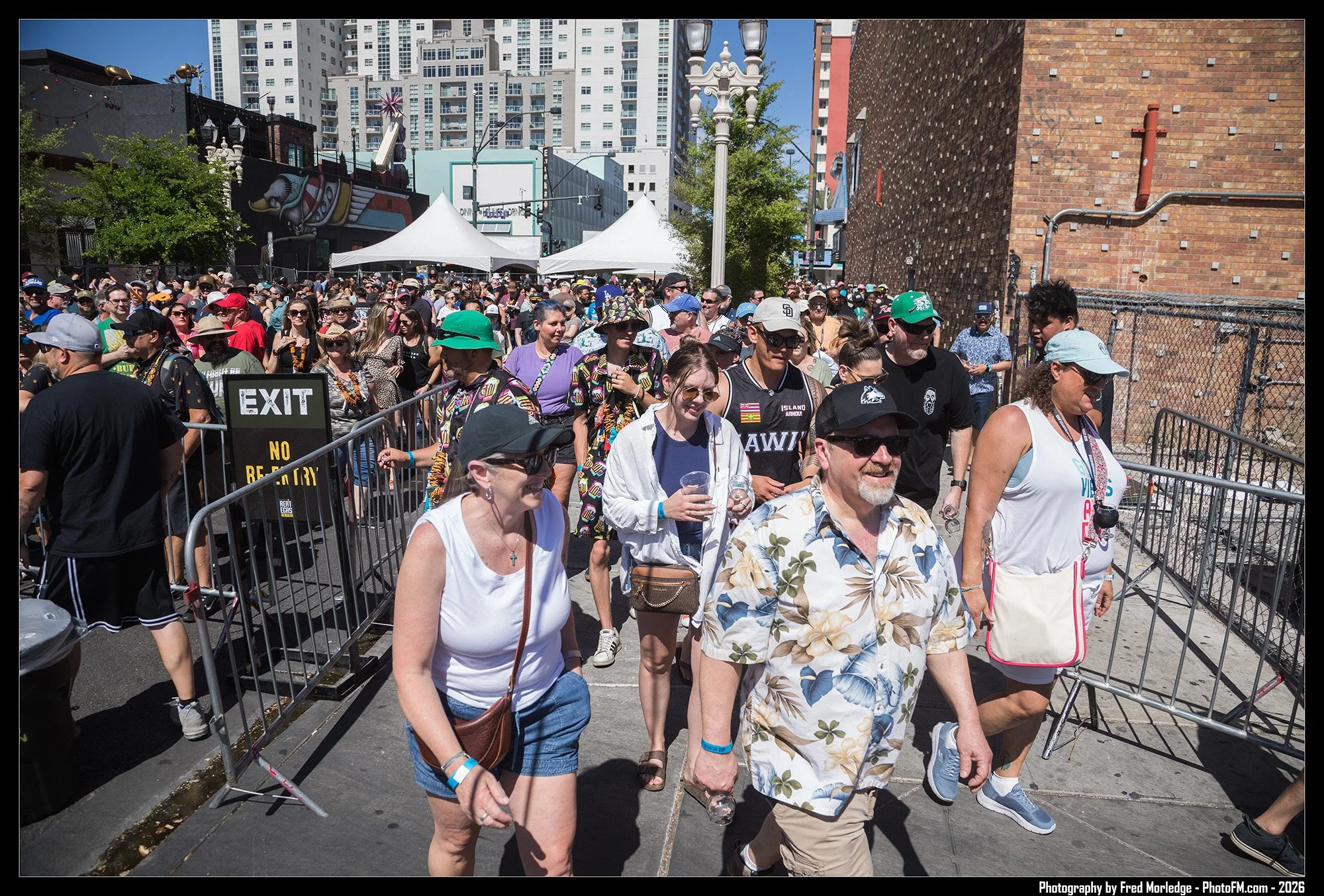 A crowd of people walking outdoors in an urban area on a sunny day, with some wearing hats, sunglasses, and casual summer clothing. The background includes tall buildings, tents, streetlights, and a brick wall.