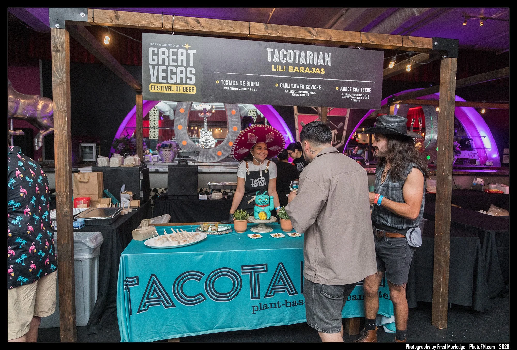 People at a taco stand with a Great Vegas Festival of Beer sign overhead, selling plant-based tacos with a woman wearing a large sombrero and a man with long hair in a sleeveless shirt interacting with customers.