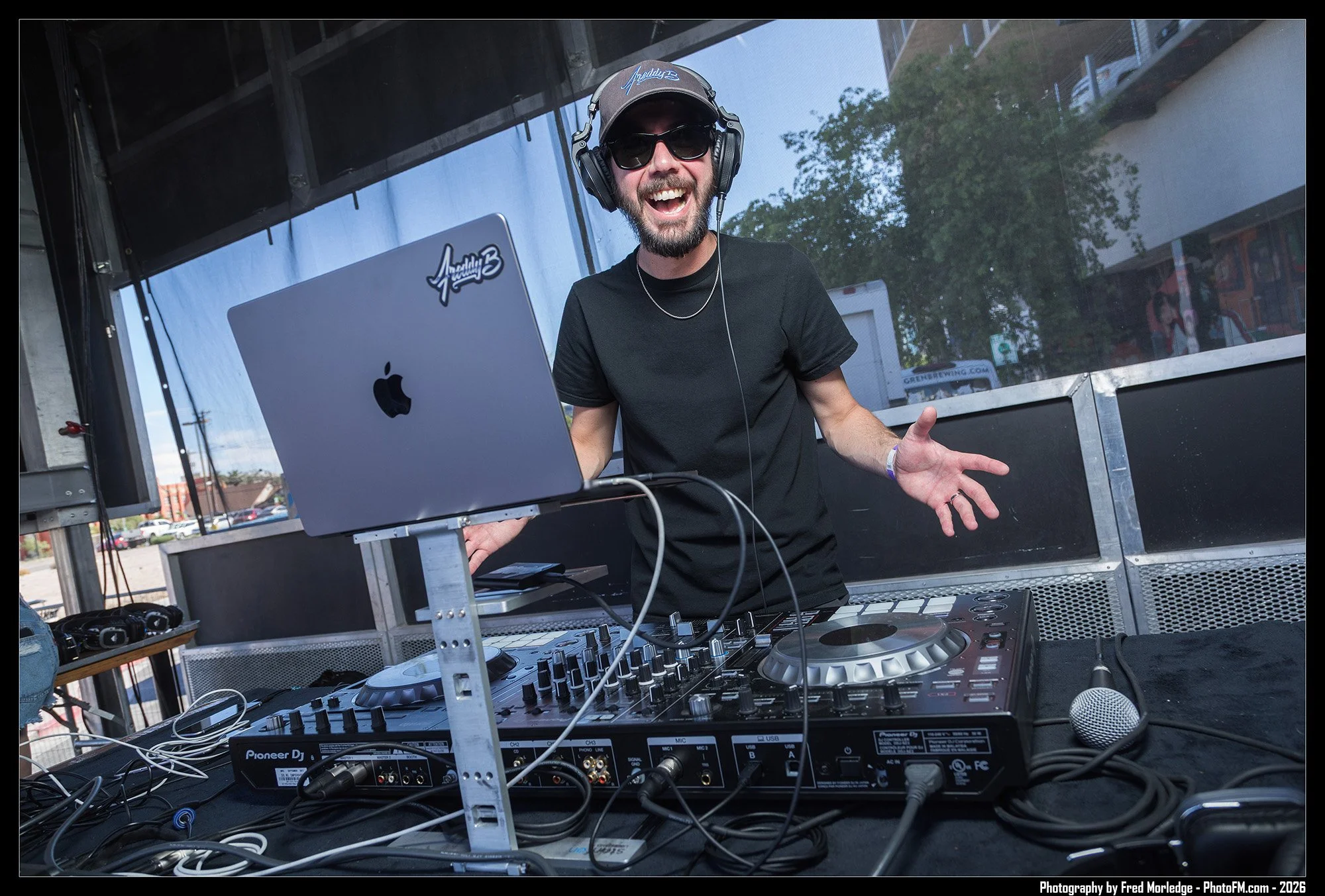 A DJ with sunglasses, headphones, and a black t-shirt, smiling and gesturing with one hand while standing behind DJ equipment, including a laptop and controllers, outdoors with buildings and trees in the background.