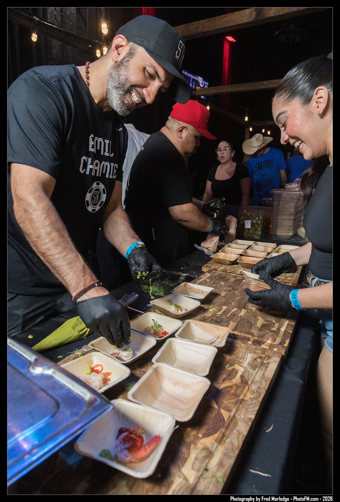 People preparing and plating sushi at a food stand or restaurant with small wooden and paper trays on a counter, using black gloves and various ingredients.