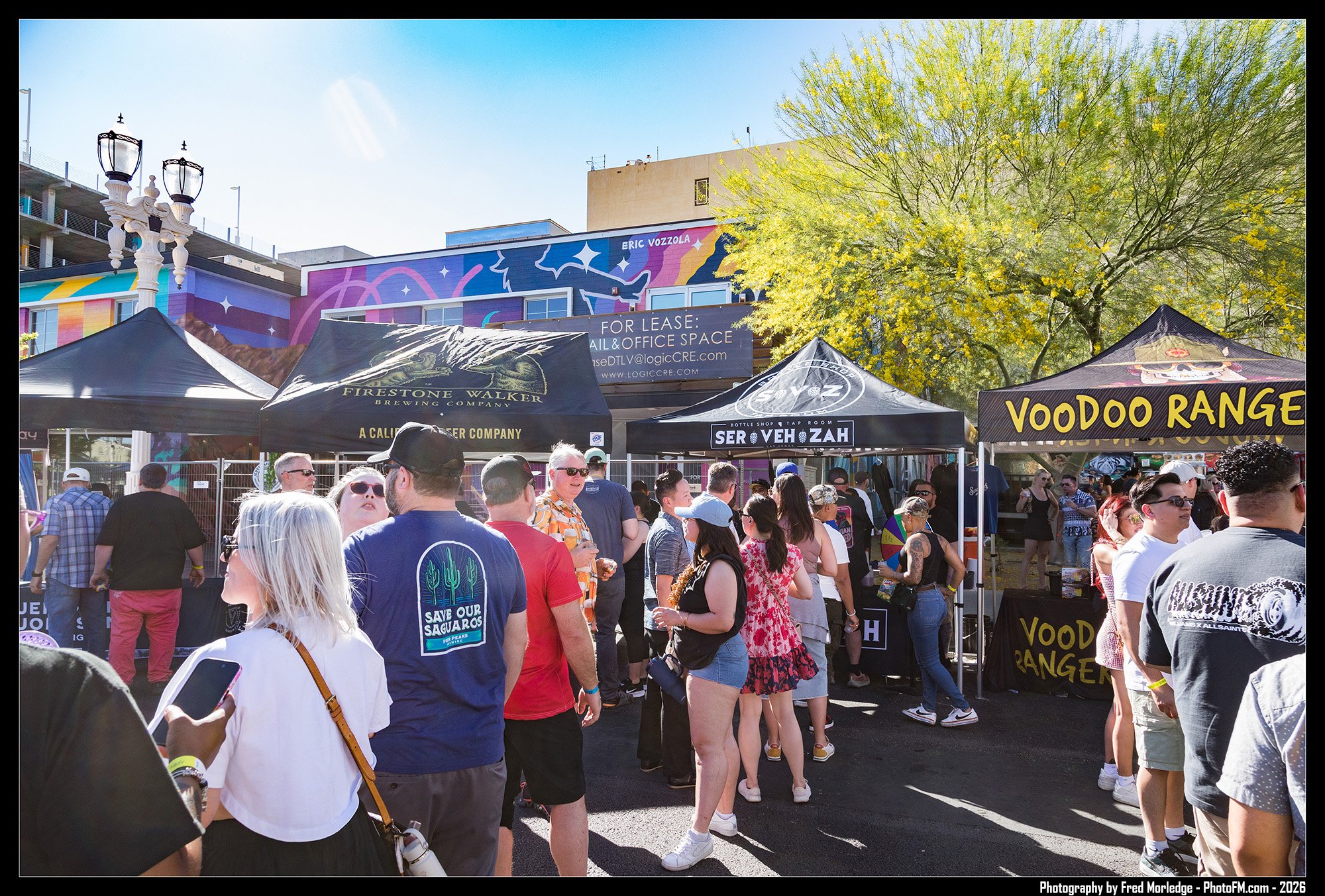 A crowd of people gathered at an outdoor event with vendor tents, trees, and colorful murals in the background on a sunny day.