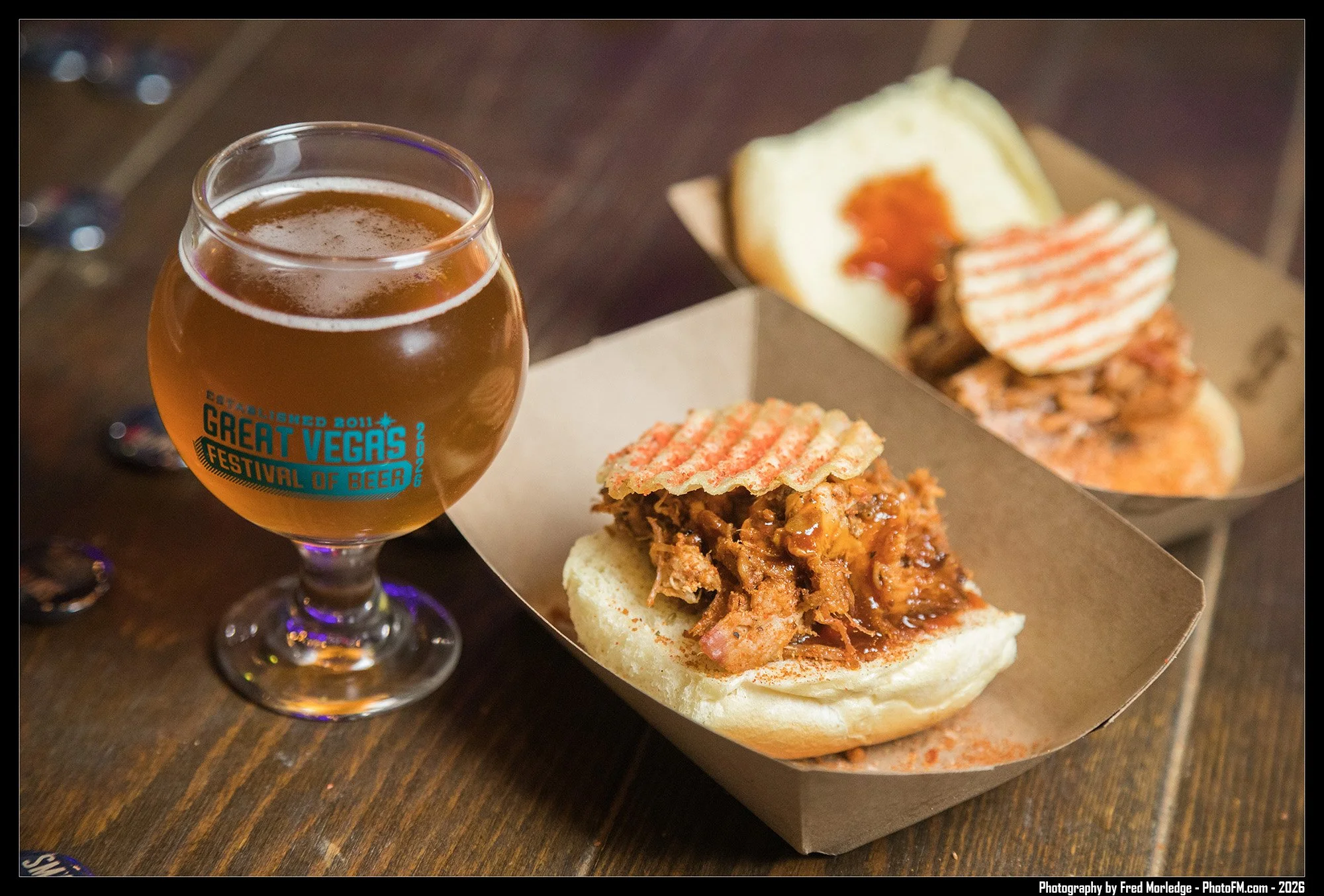 A glass of beer next to a sandwich with shredded meat, cheese, and BBQ sauce on a bun, on a wooden table.