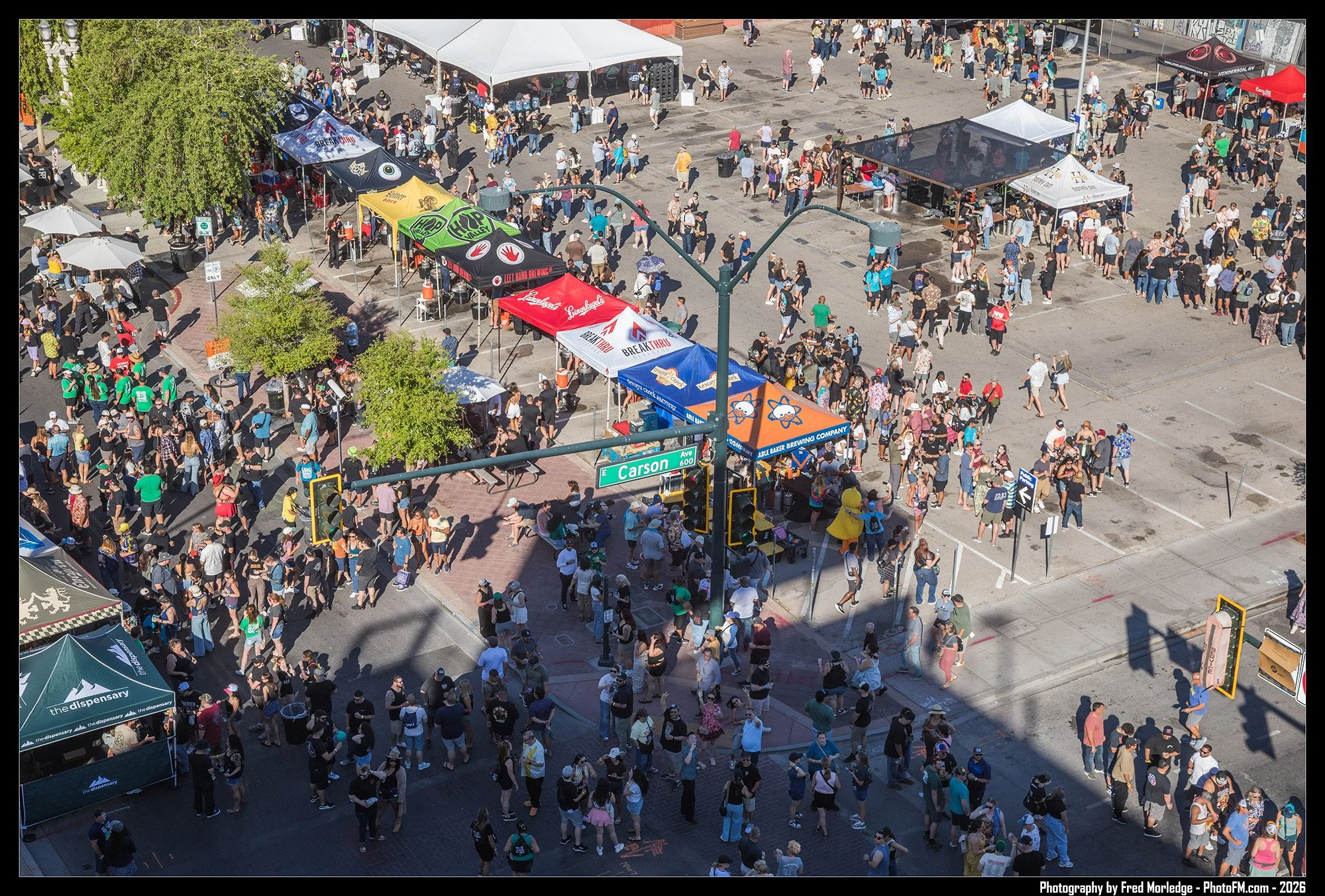 Crowd of people standing in line at an outdoor event with tents and booths on a city street.