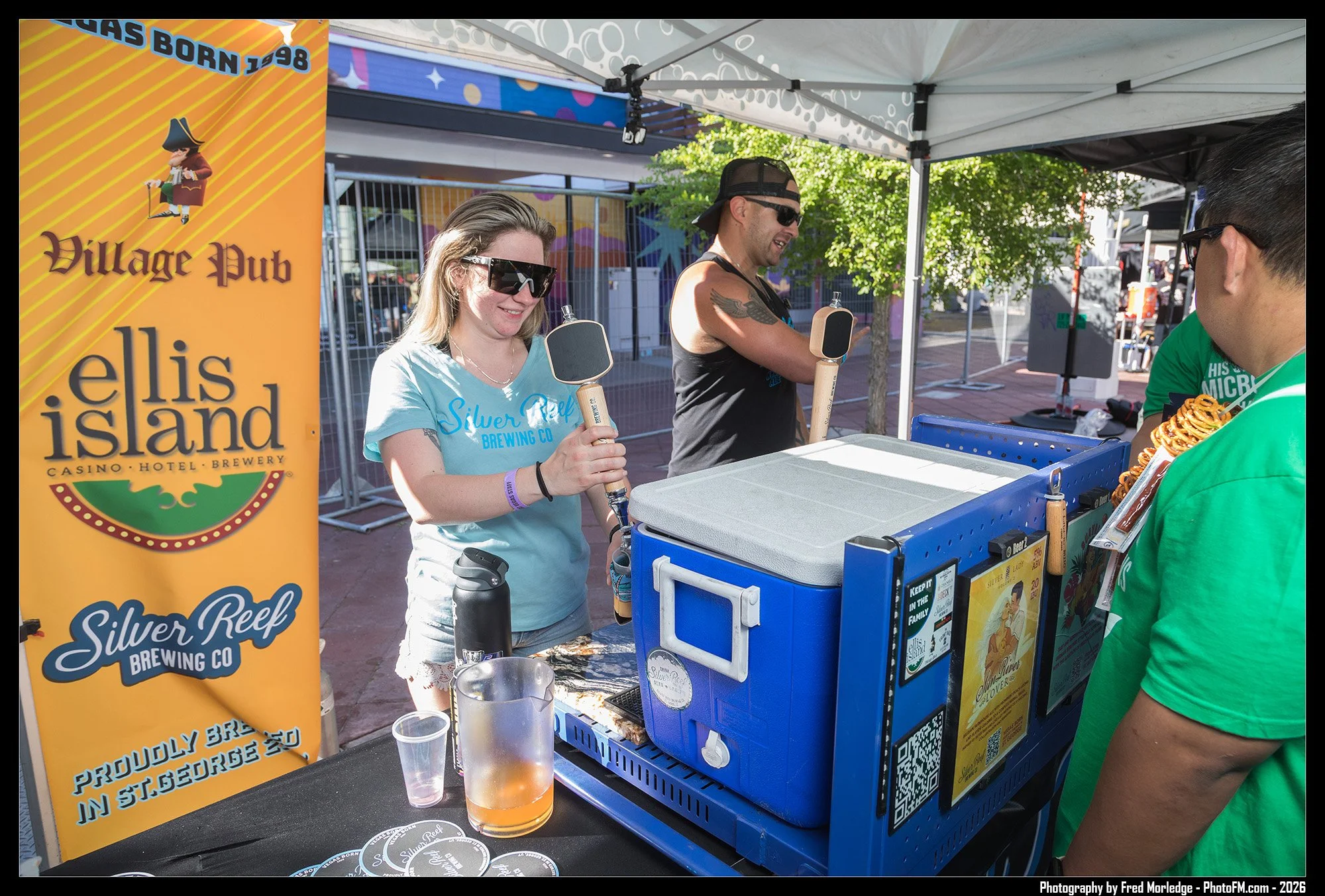 People at a booth with large beer paddles, serving drinks, at the Silver Reef Brewing Co. event, with a yellow banner and outdoor setting.