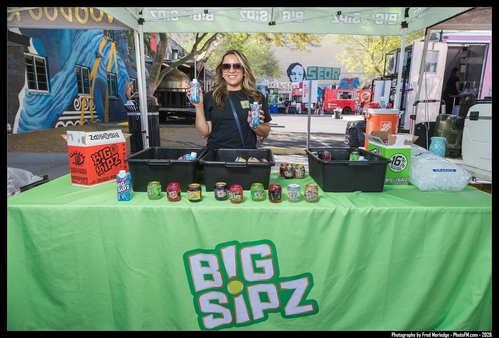 A woman wearing sunglasses and a black shirt stands behind a green-covered table at an outdoor event. She holds a can and a small box of Big Sipz drinks. The table displays several cans of Big Sipz flavored beverages, and there are empty bins and a b