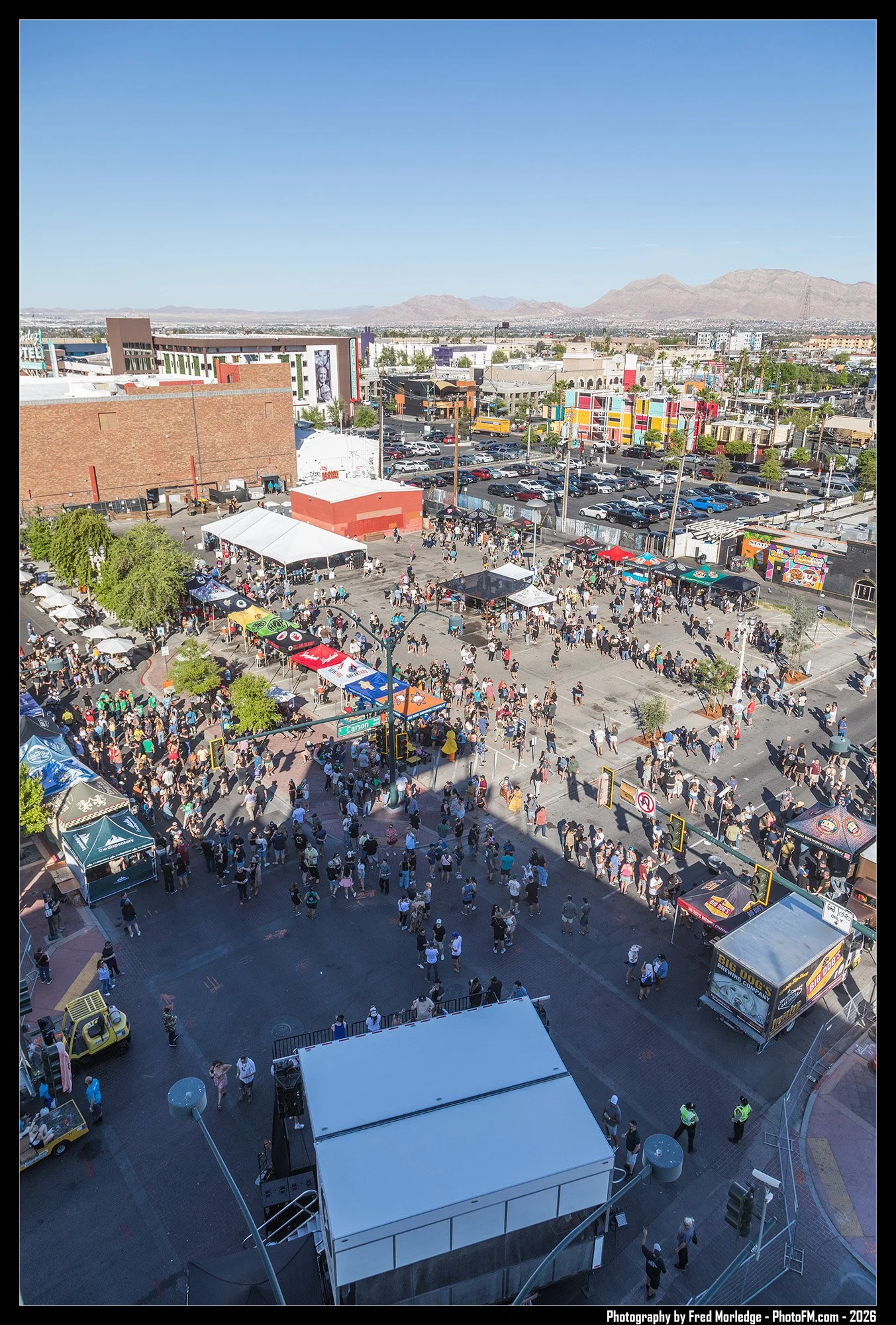 Crowd gathered at an outdoor event in a city with various tents and booths set up, street signs and mountains visible in the background.