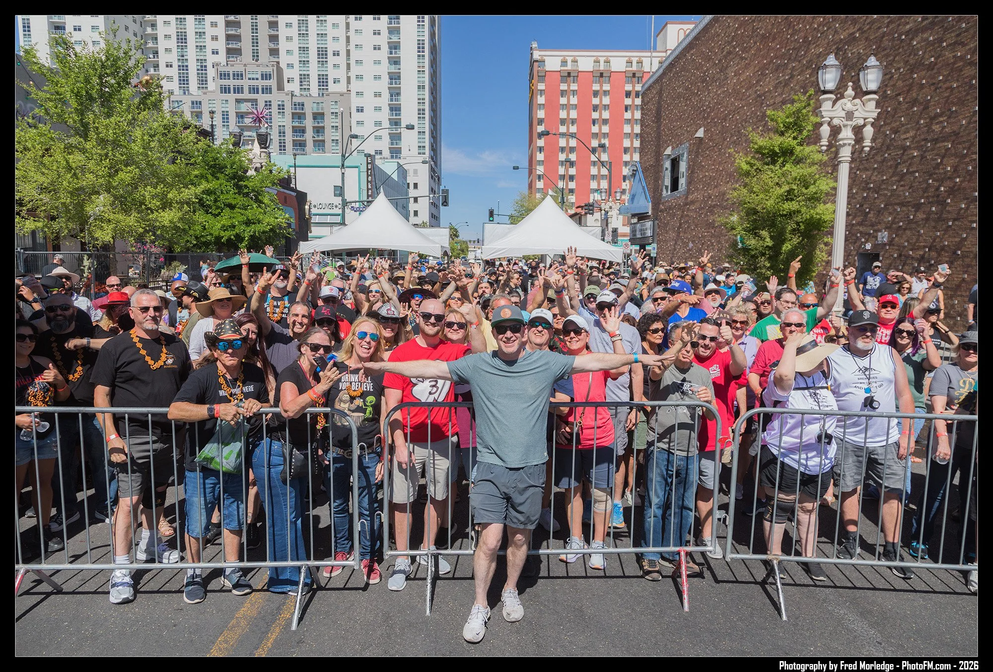 A large crowd of people gathered outdoors in front of metal barricades, with some raising their hands and smiling. Several are wearing sunglasses and casual clothing, and many are holding drinks or smartphones. The scene is sunny with a clear blue sk
