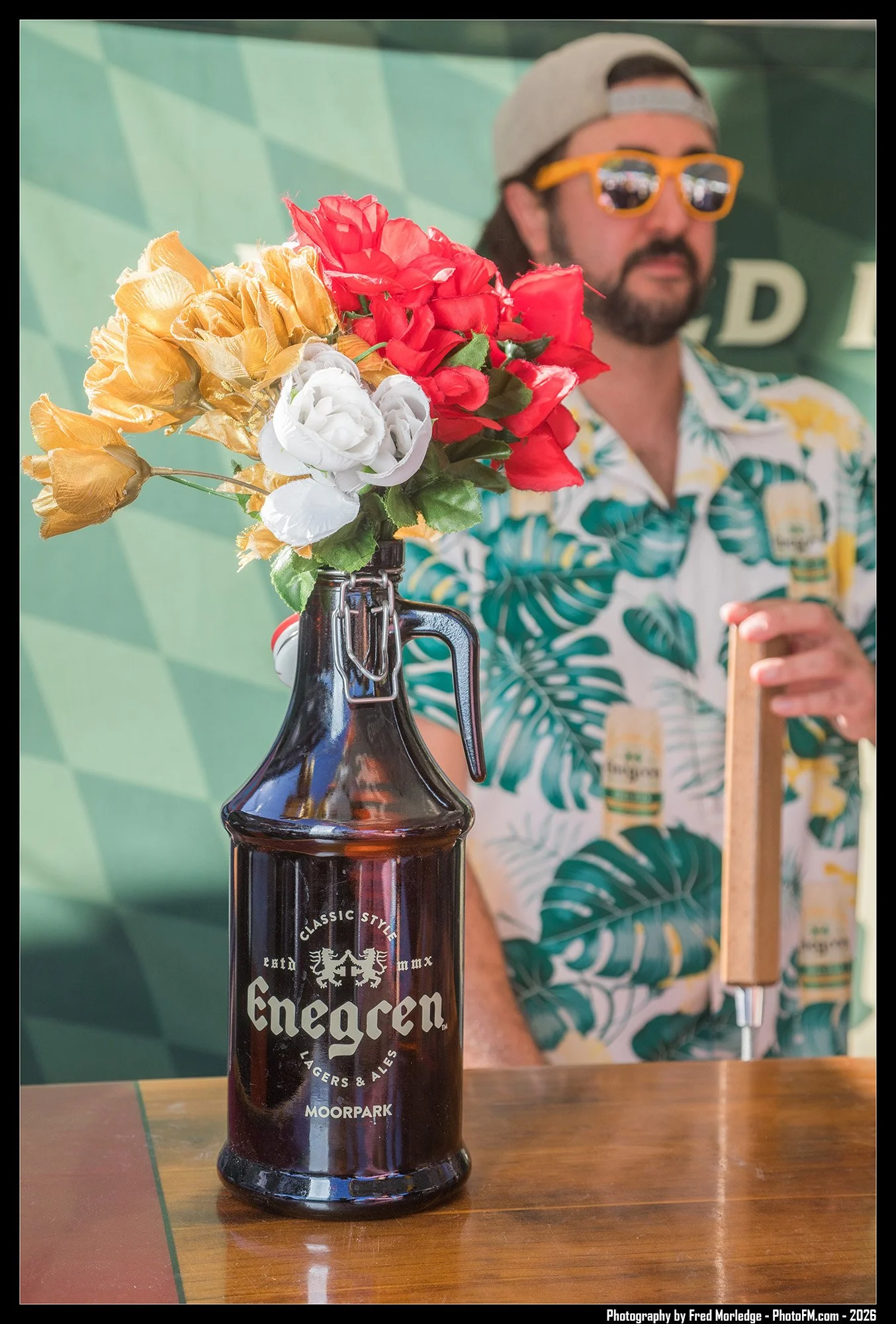A dark glass bottle of Enegren beer with a colorful artificial flower arrangement attached to its neck, placed on a wooden table. In the background, a man wearing a patterned Hawaiian shirt, sunglasses, and a gray cap is standing near a green wall.