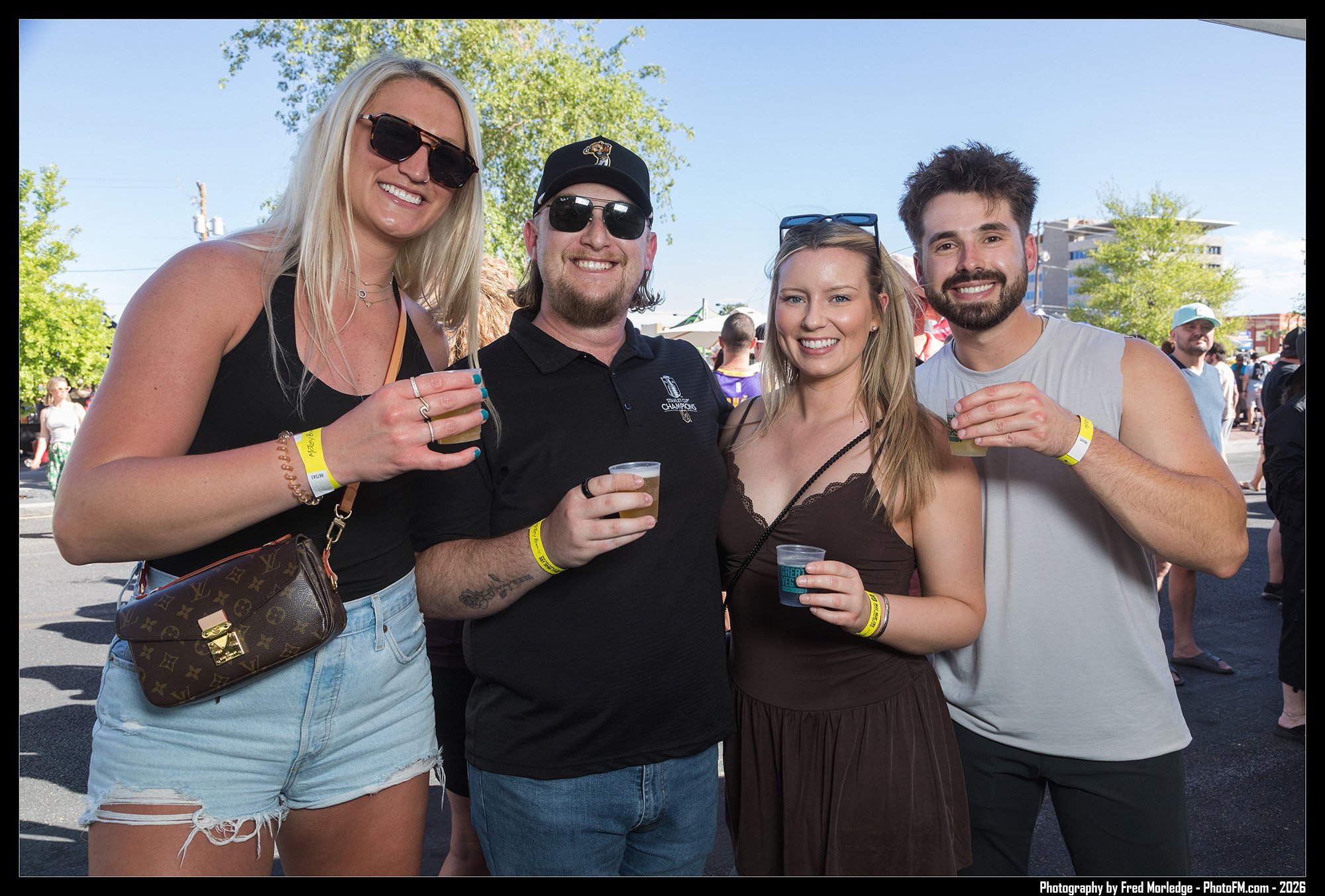 Four people standing outdoors on a sunny day at a crowd event, smiling and holding drinks, with trees and buildings in the background.