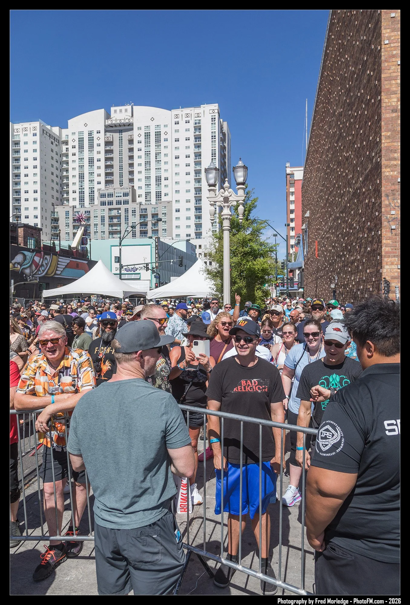 A crowd of people gathered outdoors under a clear blue sky, with some behind a metal barricade. In the background, there are tall modern white apartment buildings, a brick building on the right, and street lamps. Some people are smiling and taking ph