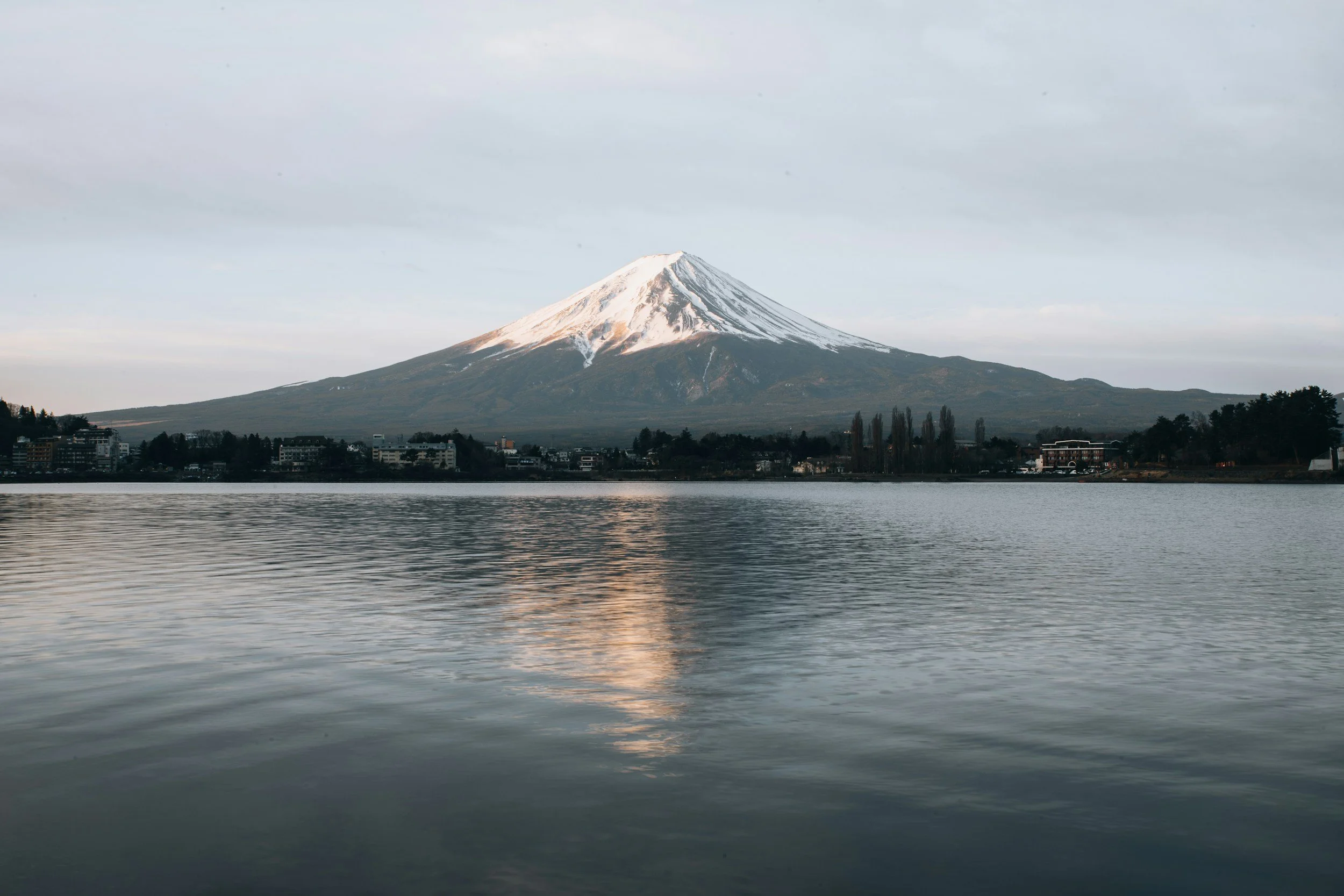 Snow-capped mountain, likely a volcano, seen across a body of water with reflections, with a cityscape and trees in the foreground under a cloudy sky.