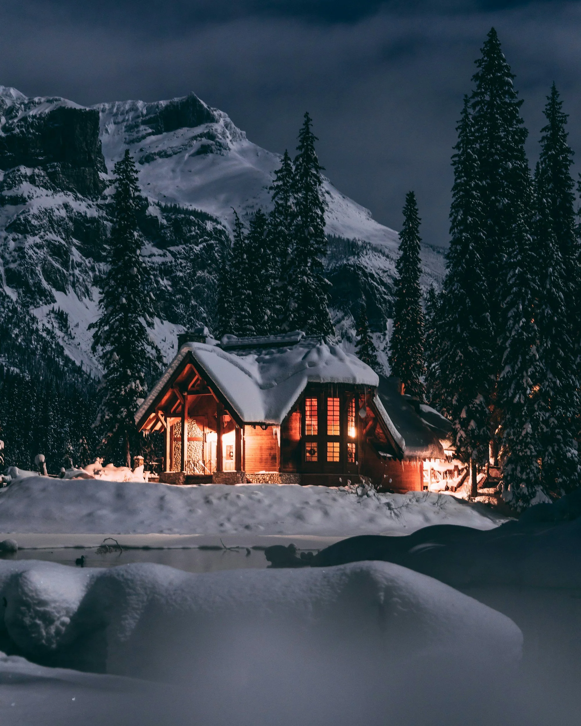 Snow-covered mountain landscape at night with a cozy lit cabin in the foreground and tall evergreen trees.