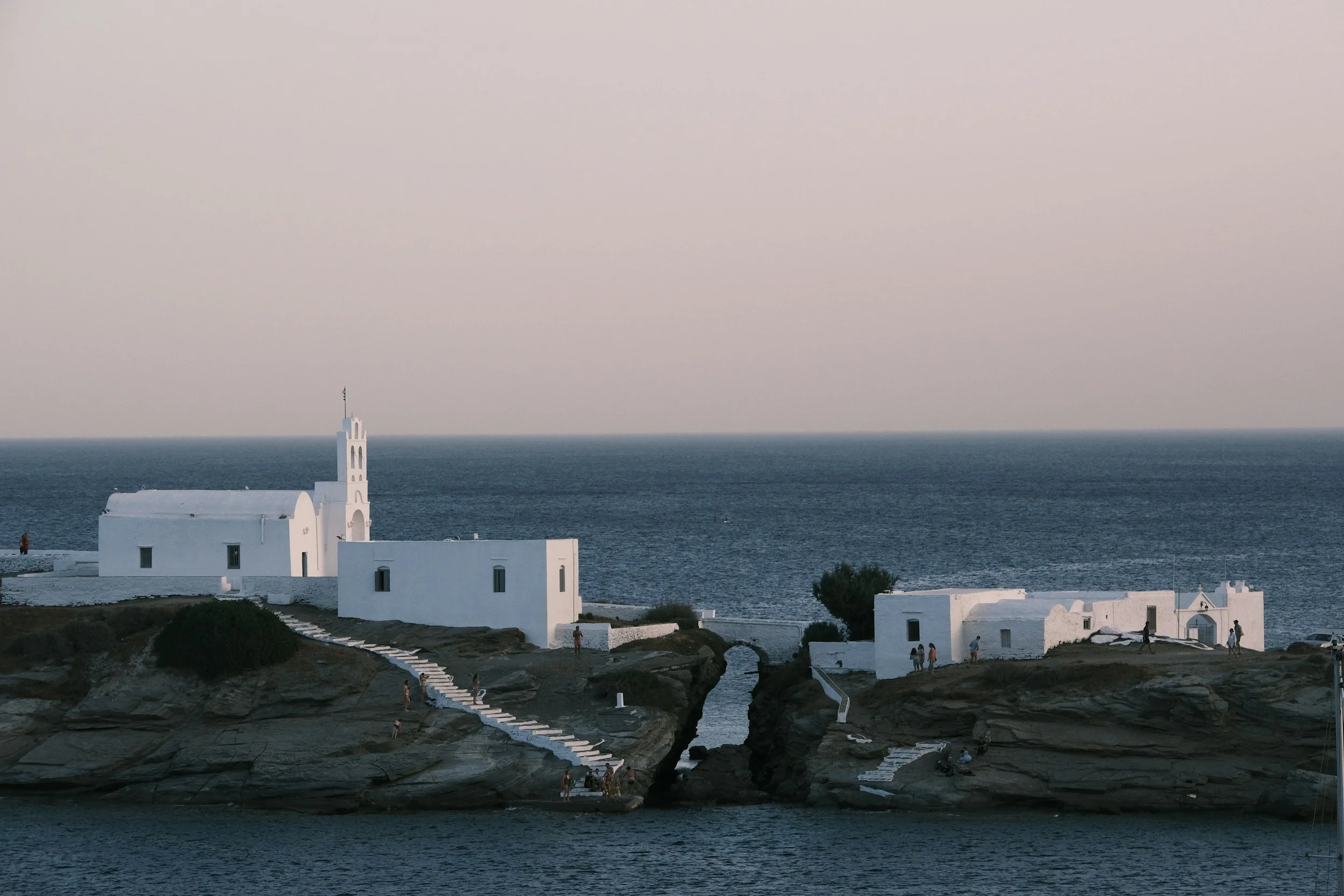White buildings with a small church in the foreground on a seaside cliff, with a staircase connecting the buildings to the water, and a cloudy sky in the background.