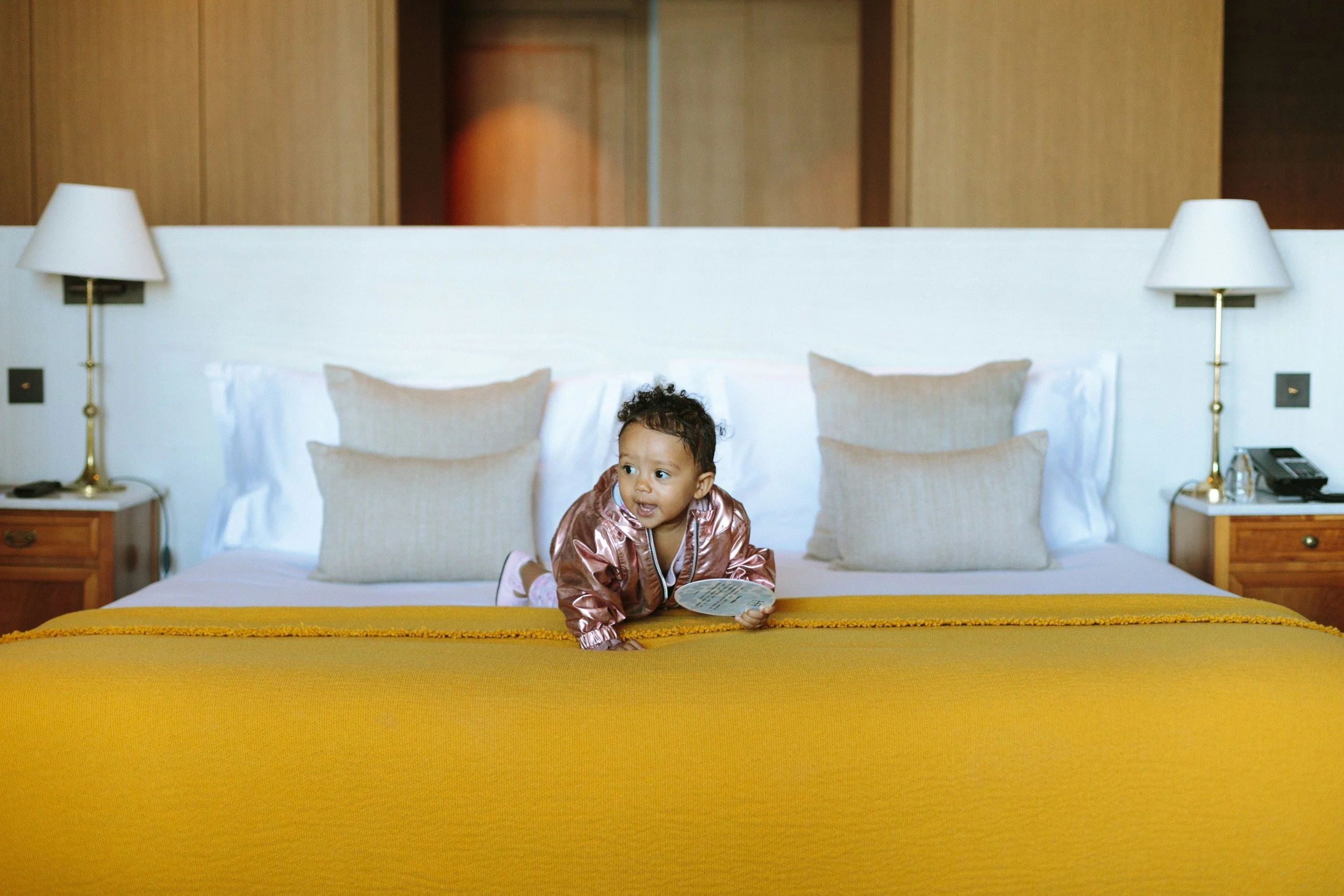 A young child with curly hair wearing a shiny pink jacket, sitting on a bed with a yellow bedspread, holding and looking at a fan of paper or cards, in a hotel room with beige pillows, lamps on bedside tables, and a wooden wall.